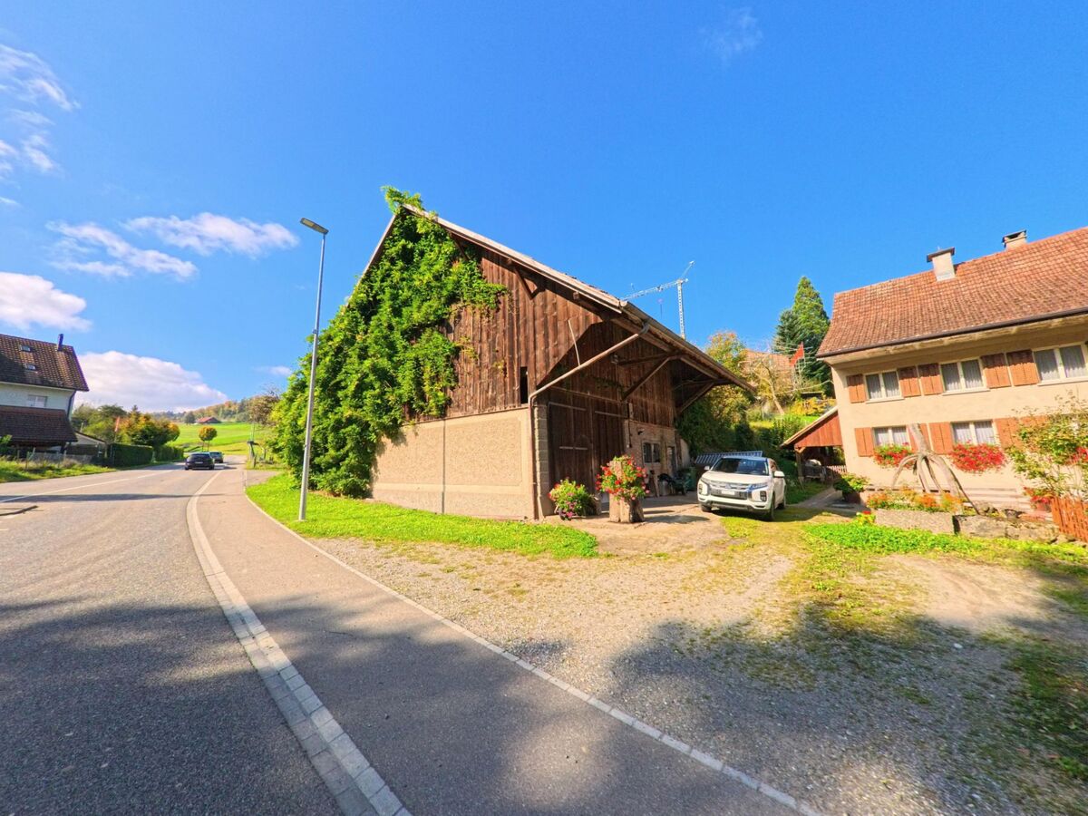 Am Rand einer leicht ansteigenden Strasse in Siglistorf (5462, Kanton Aargau) stehen ein traditioneller Bauernhof mit Rankpflanzen und ein Wohnhaus mit Blumenschmuck.