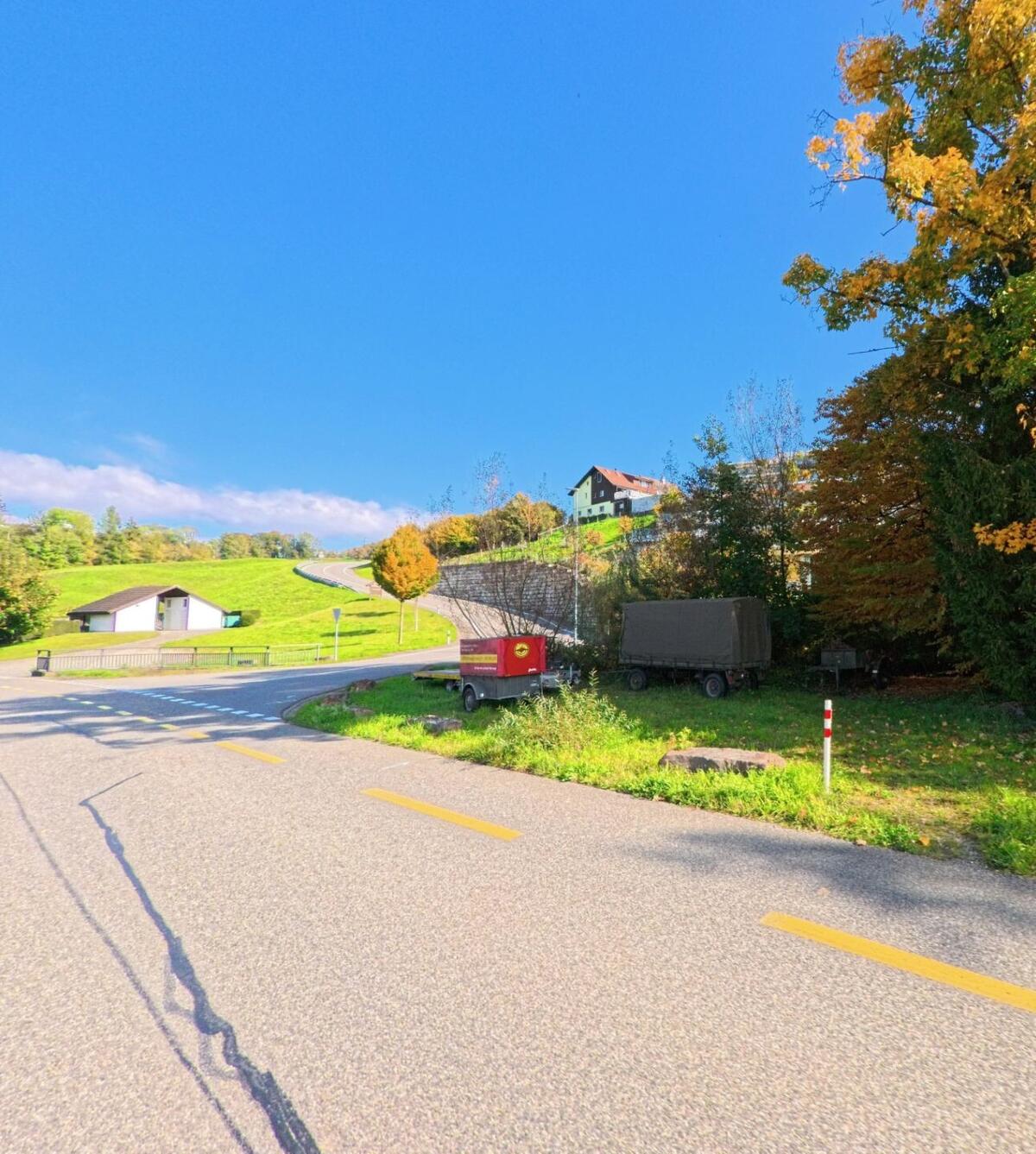 An einer Kurve in Siglistorf (5462, Kanton Aargau) fuehrt die Strasse durch eine gruene Huegellandschaft, waehrend zwei Anhaenger am Waldrand abgestellt sind.