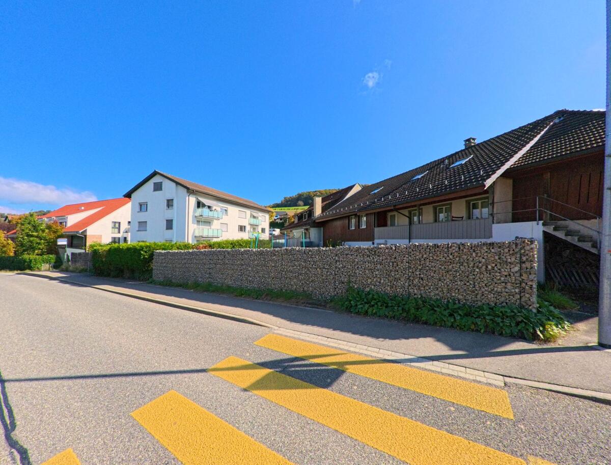 Entlang einer Strasse in Siglistorf (5462, Kanton Aargau) stehen moderne und traditionelle Wohnhaeuser hinter einer hohen Steinmauer, waehrend ein Fussgaengerstreifen den Vordergrund praegt.