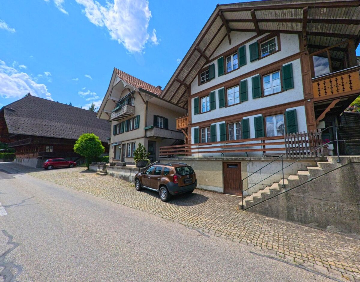 Zwei gepflegte Wohnhaeuser mit gruenen Fensterlaeden stehen in Signau (3534, Kanton Bern) nebeneinander an einer gepflasterten Strasse mit abgestellten Fahrzeugen.
