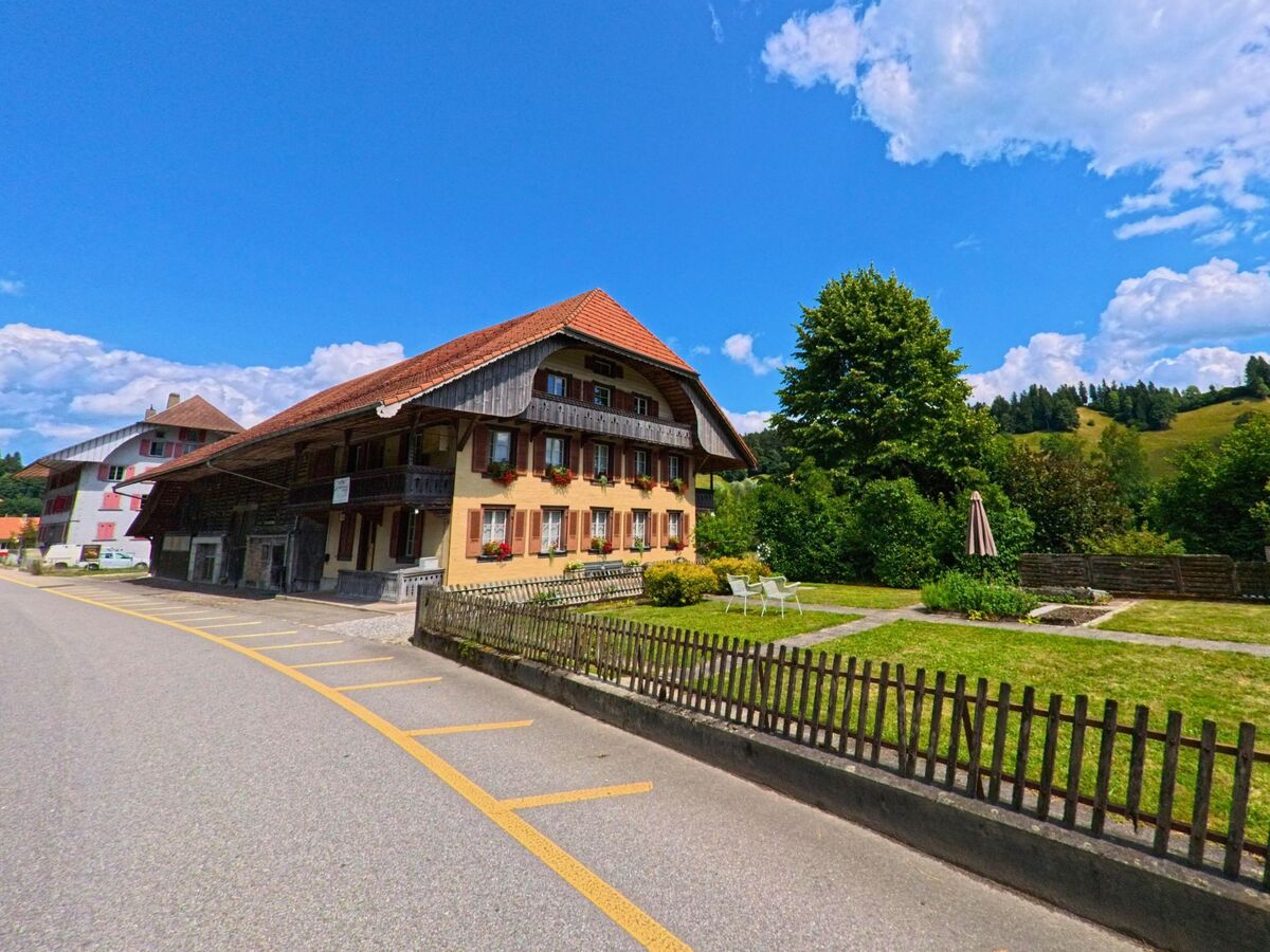 In Signau (3534, Kanton Bern) steht ein traditionelles Wohnhaus mit rotem Ziegeldach an einer Strasse, daneben ein gepflegter Garten mit Sitzgelegenheiten und Blick auf bewaldete Huegel.