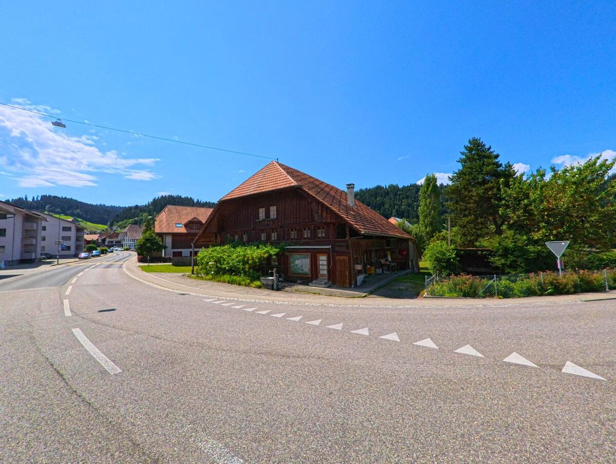 An einer verkehrsreichen Kurve in Signau (3535, Kanton Bern) steht ein traditionelles Holzhaus mit rotem Ziegeldach, umgeben von Wohnhaeusern und gruener Vegetation.