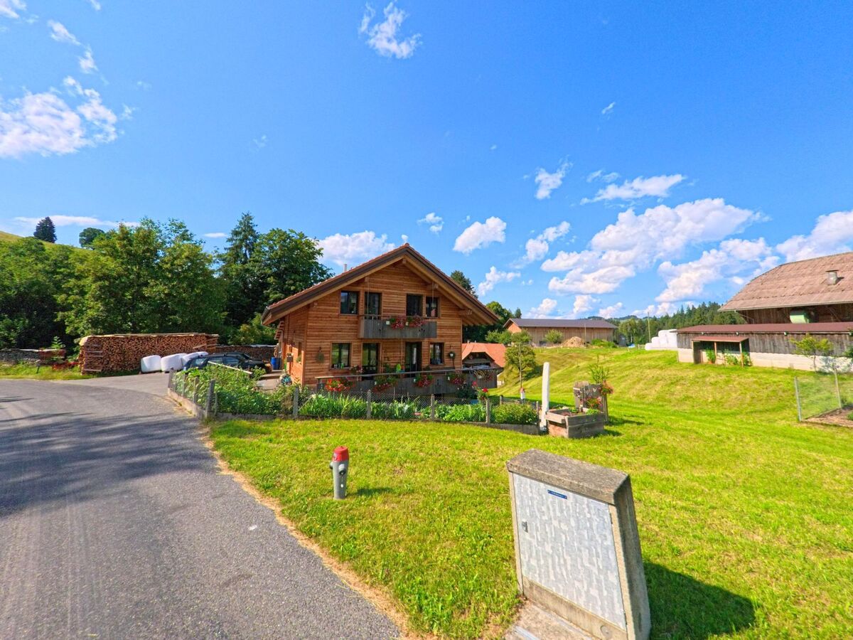 Ein Holzhaus mit Blumenschmuck steht in Sigriswil (3657, Kanton Bern) neben einer schmalen Landstrasse, umgeben von Gruenflaechen und Bauerngebaeuden.