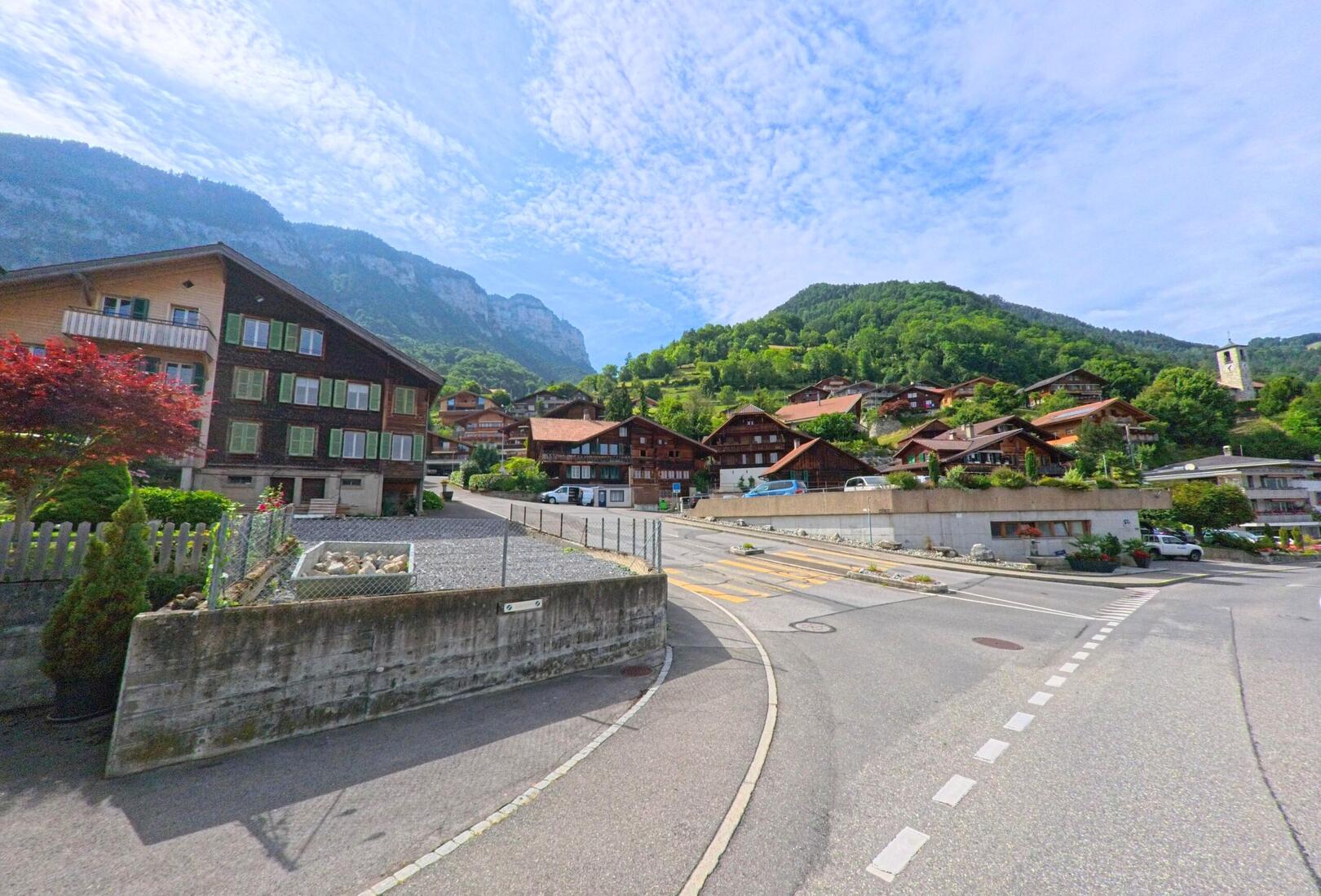 In Sigriswil (3658, Kanton Bern) steigt die Dorfstrasse durch eine Hanglage mit zahlreichen Chalets an, im Hintergrund erhebt sich eine markante Felswand und ein Kirchturm ist sichtbar.