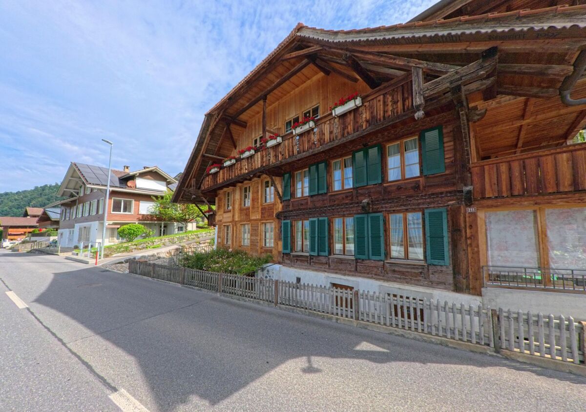 Ein traditionelles Holzhaus mit gruenen Fensterlaeden steht in Sigriswil (3658, Kanton Bern) direkt an einer leicht abschuessigen Dorfstrasse, daneben moderne Wohnhaeuser.