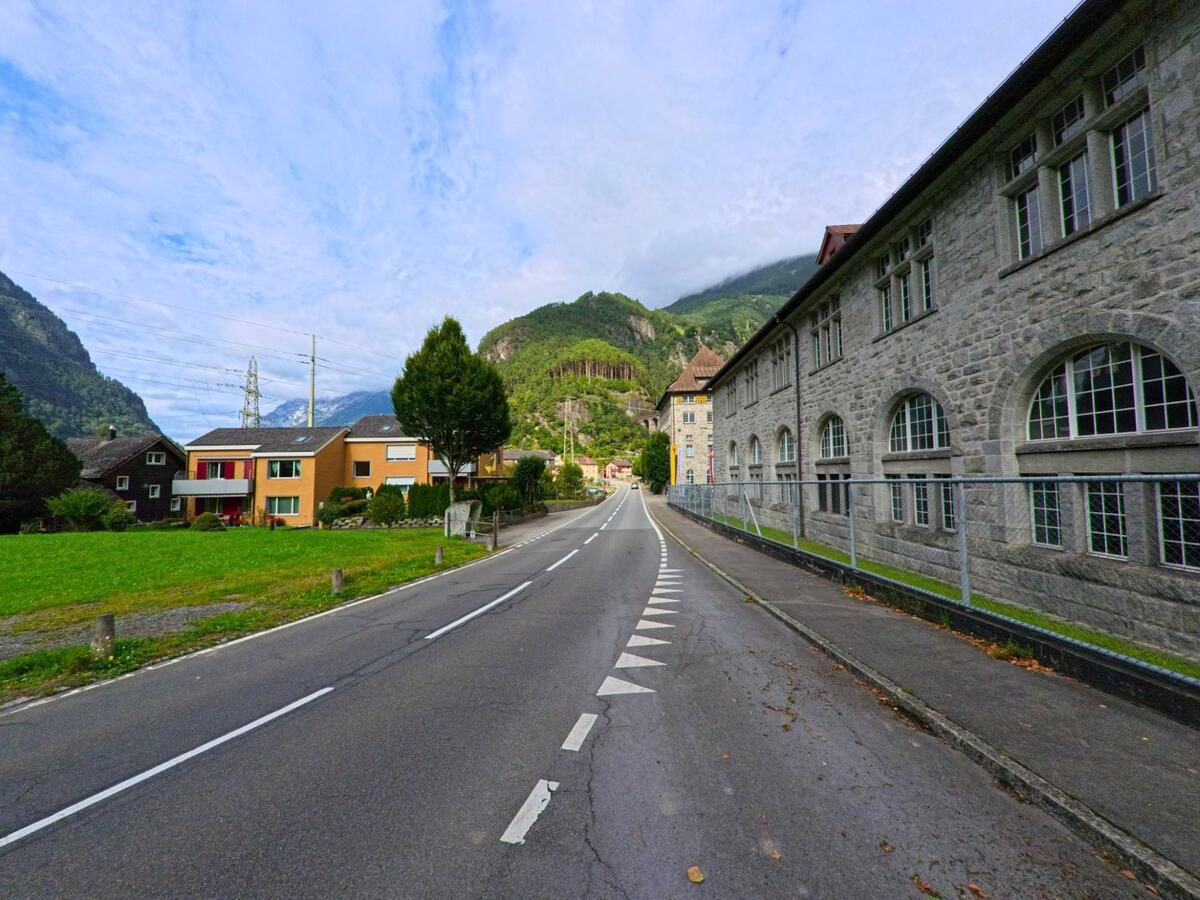 Eine Hauptstrasse fuehrt in Silenen (6474, Kanton Uri) zwischen einem grossen Steingebaeude und modernen Wohnhaeusern in Richtung der bewaldeten Berghaenge.