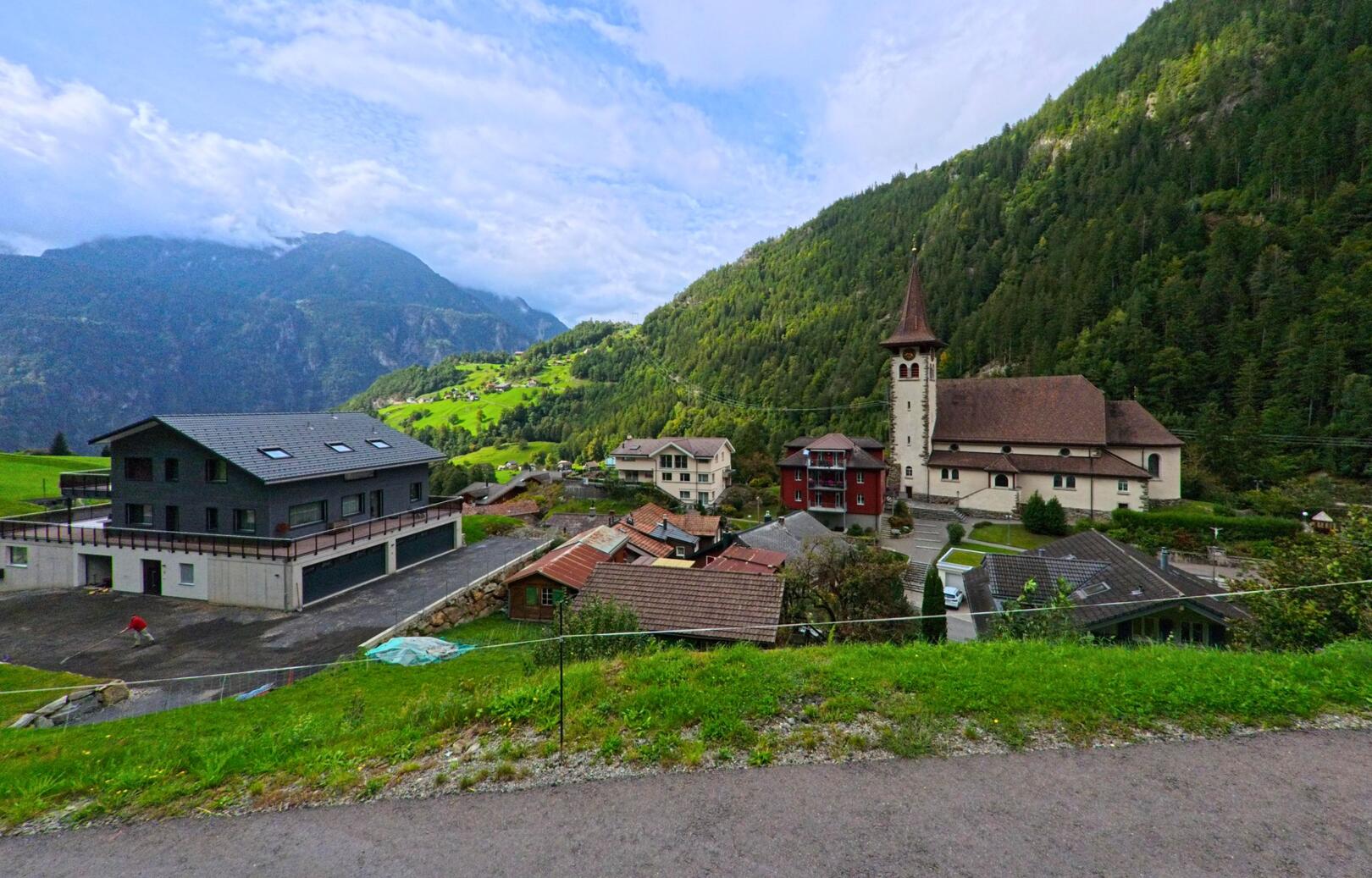 In Silenen (6475, Kanton Uri) liegt die Pfarrkirche mit ihrem markanten Turm eingebettet zwischen Wohnhaeusern und bewaldeten Berghaengen unter einem teilweise wolkigen Himmel.