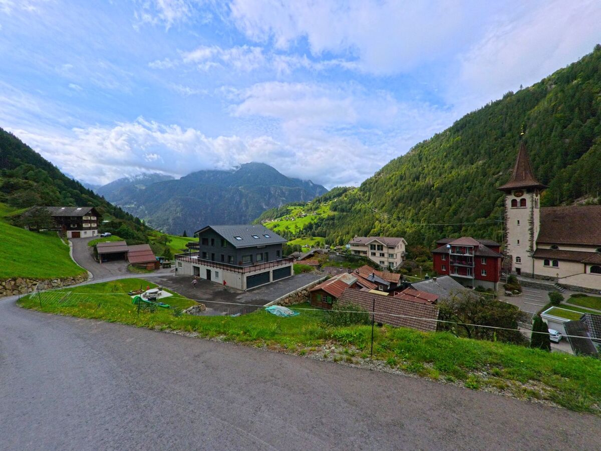 Von einer ansteigenden Strasse aus bietet sich in Silenen (6475, Kanton Uri) ein weiter Blick auf die Dorfkirche, Wohnhaeuser und die umliegenden bewaldeten Berghaenge.