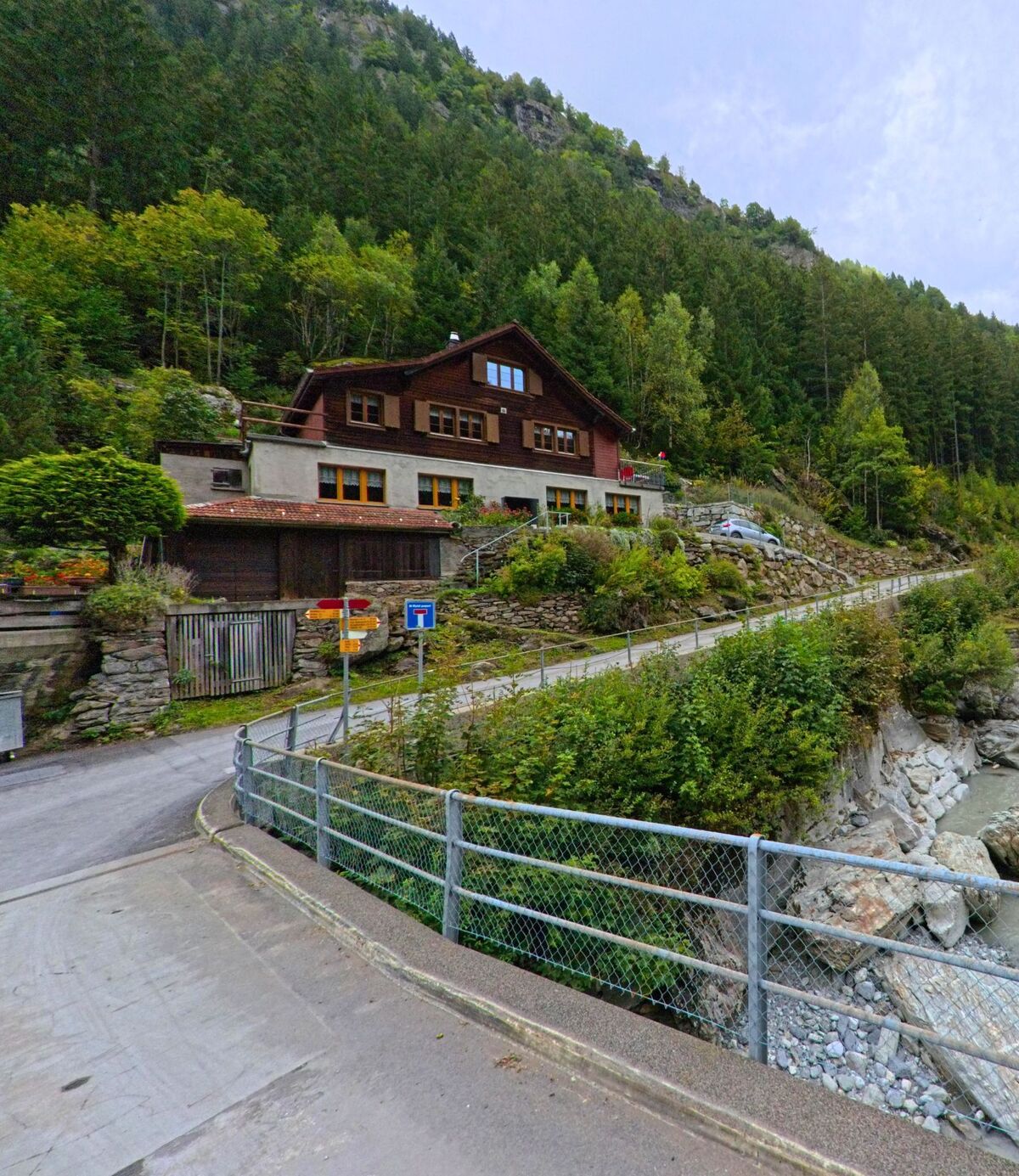 Ein Holzhaus mit Steinfundament liegt in Silenen (6475, Kanton Uri) oberhalb eines Baches am Waldrand, umgeben von dichter Vegetation und steilen Felswaenden.