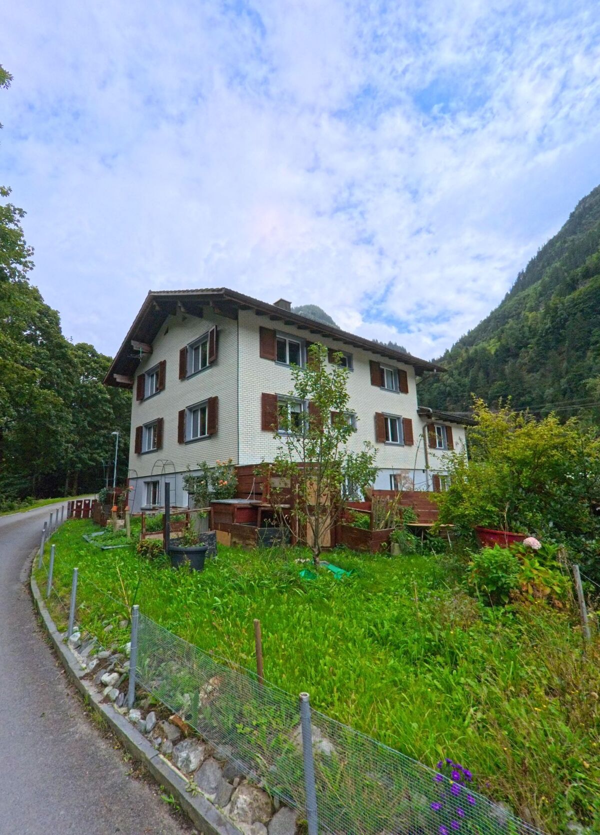 Ein mehrstöckiges Wohnhaus mit braunen Fensterlaeden steht in Silenen (6475, Kanton Uri) an einer schmalen Strasse, umgeben von Gruenflaechen und dichtem Bergwald.