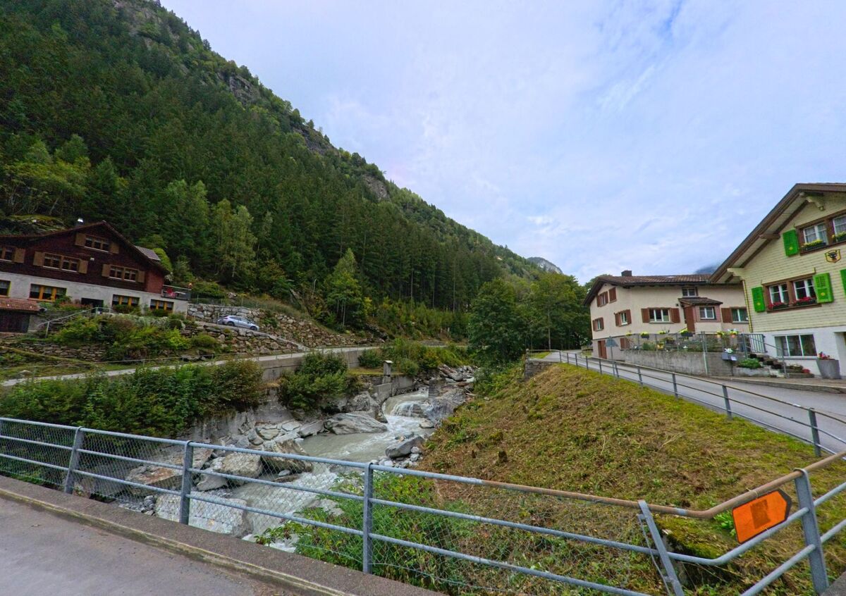 In Silenen (6475, Kanton Uri) verlaeuft eine schmale Strasse entlang eines Gebirgsbaches, flankiert von Wohnhaeusern und dichten Nadelwaeldern am steilen Talhang.
