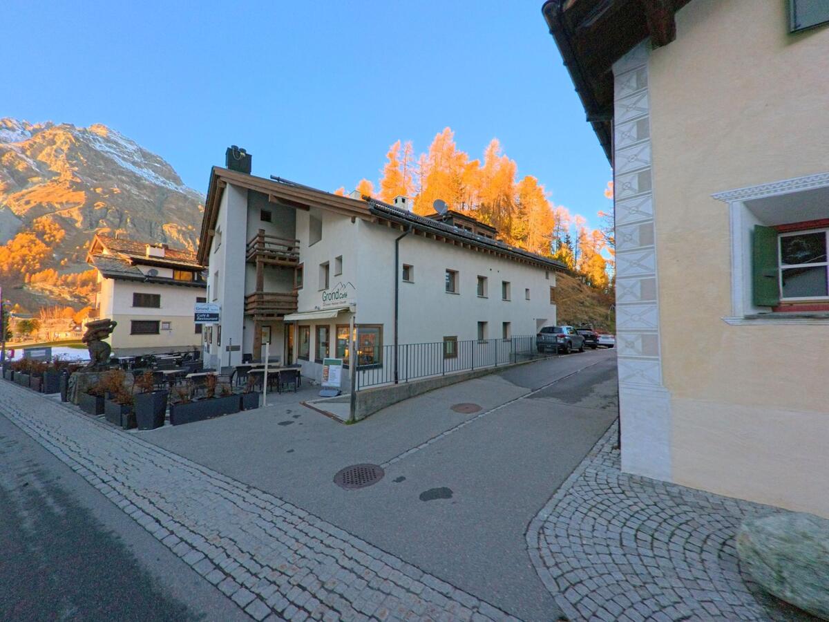 An einer kleinen Verzweigung mit Cafeterrasse und Wohngebäuden zeigt sich eine ruhige Dorfzone in Sils im Engadin/Segl (7514, Kanton Graubünden), umrahmt von herbstlich leuchtenden Lärchenhängen.