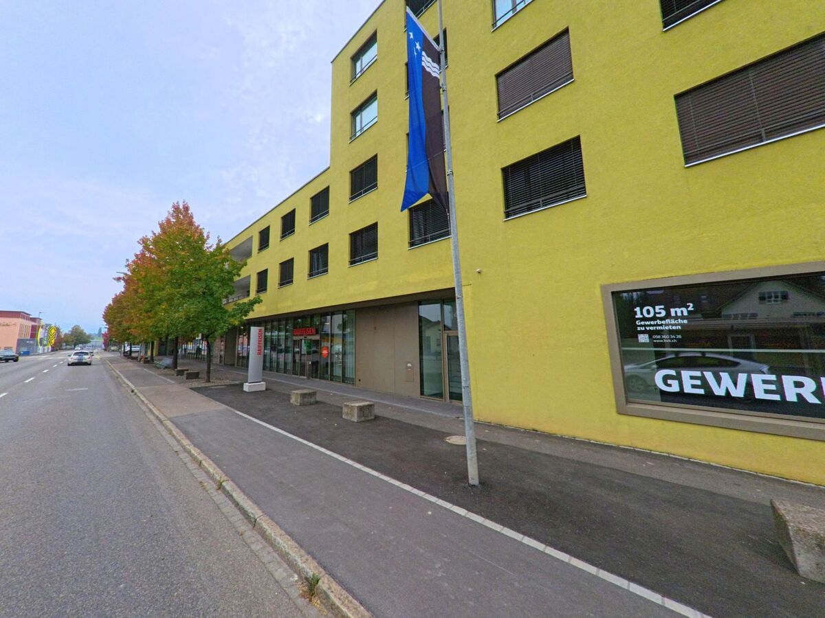 Entlang der Hauptstrasse in Sins (5643, Kanton Aargau) steht ein modernes gelbes Gebaeude mit Gewerbeflaechen, vor dem herbstlich gefaerbte Baeume und ein Gehweg verlaufen.