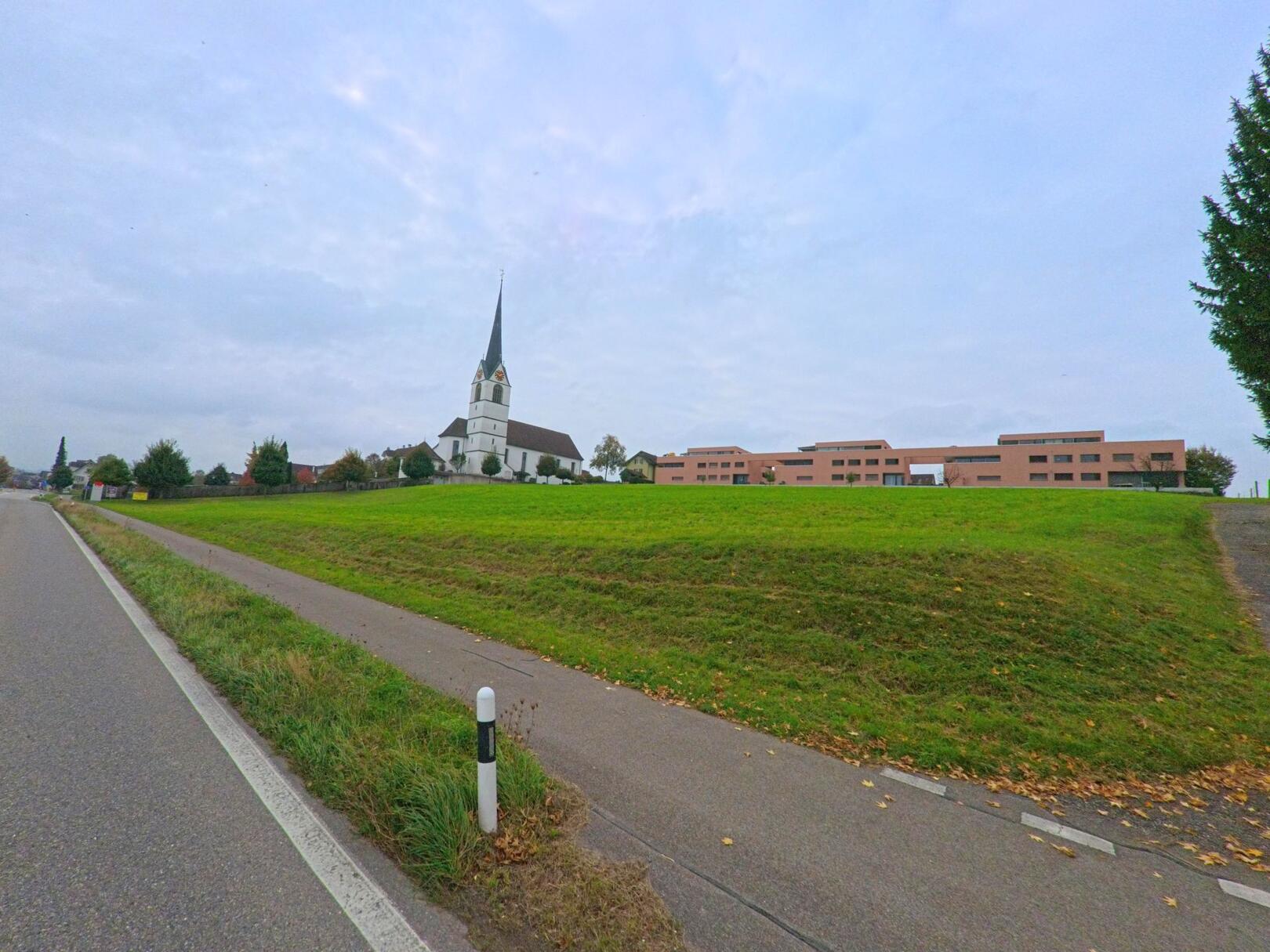 Auf einer leicht ansteigenden Wiese in Sins (5643, Kanton Aargau) steht die Kirche gut sichtbar neben modernen Gebäuden, während der Weg entlang der Strasse vorbeiführt.