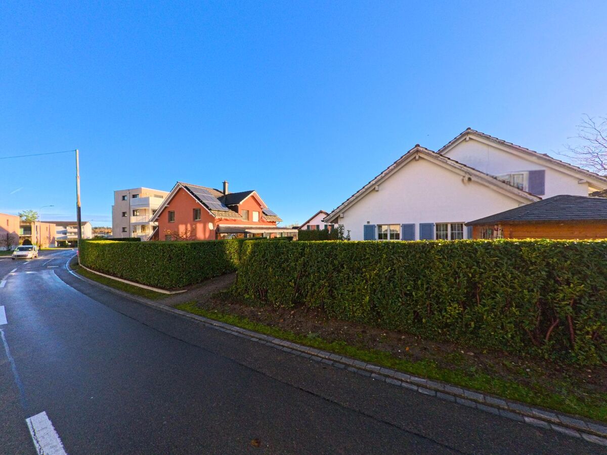 Entlang einer leicht geschwungenen Quartierstrasse in Sirnach (8371, Kanton Thurgau) reihen sich Einfamilienhaeuser hinter gepflegten Hecken unter klarem Himmel.