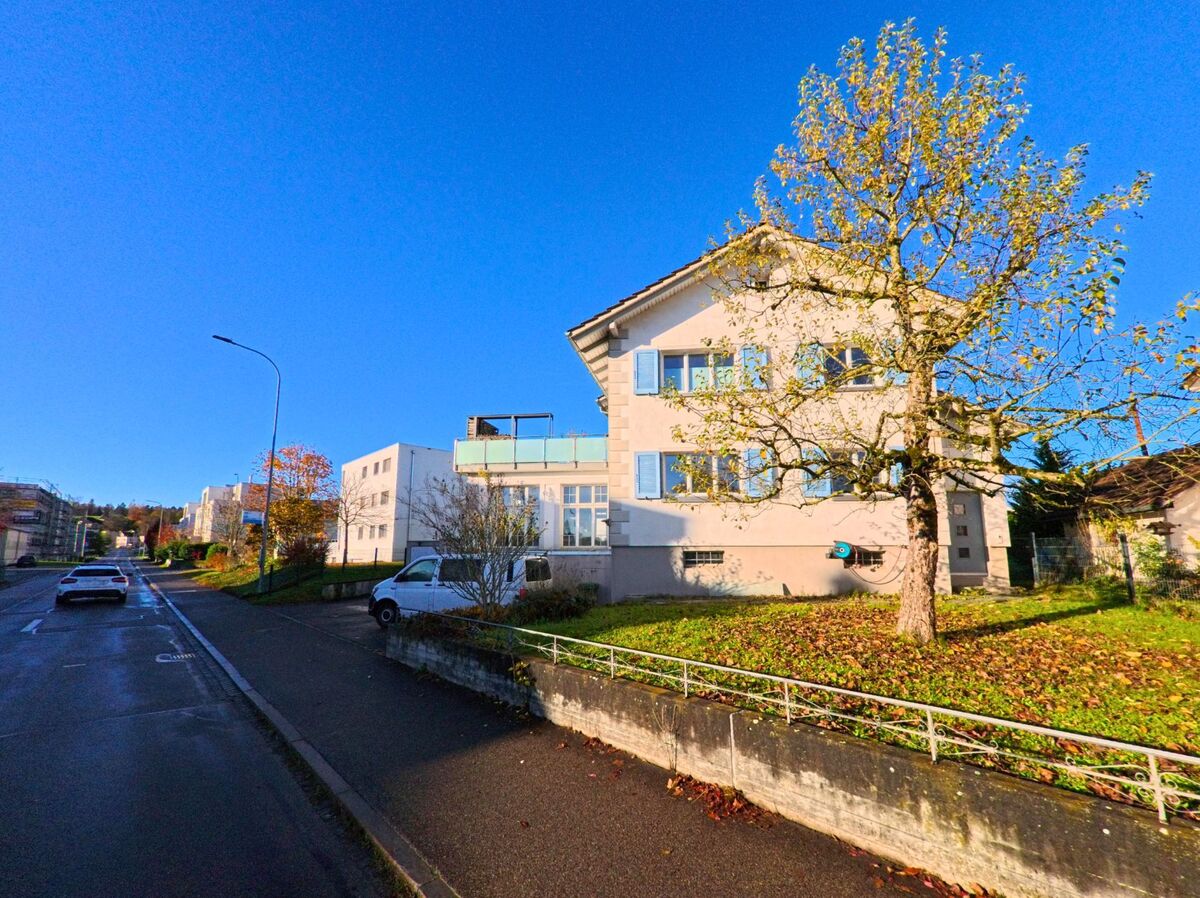 An einer leicht ansteigenden Strasse in Sirnach (8371, Kanton Thurgau) verlaufen Gehweg und Wohnbauten entlang einer Gruenflaeche mit Baum im herbstlichen Licht.