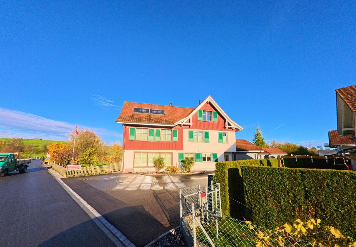 Ein traditionelles Wohnhaus mit roter Fassade und gruenen Fensterlaeden steht an einer Nebenstrasse in Sirnach (8371, Kanton Thurgau) vor offenem Himmel und herbstlicher Umgebung.