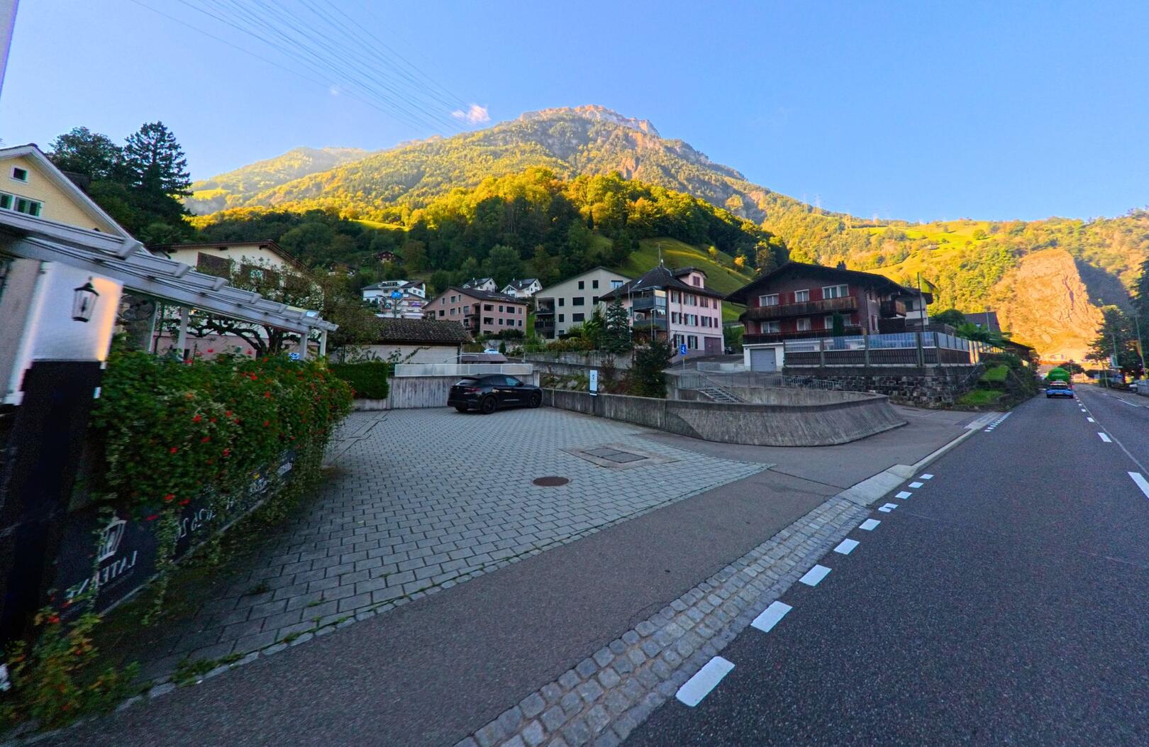 Am Rand einer ruhigen Strasse zeigt sich das Dorfgebiet von Sisikon (6452, Kanton Uri) mit Wohnhäusern vor einem sonnenbeschienenen Berghang.