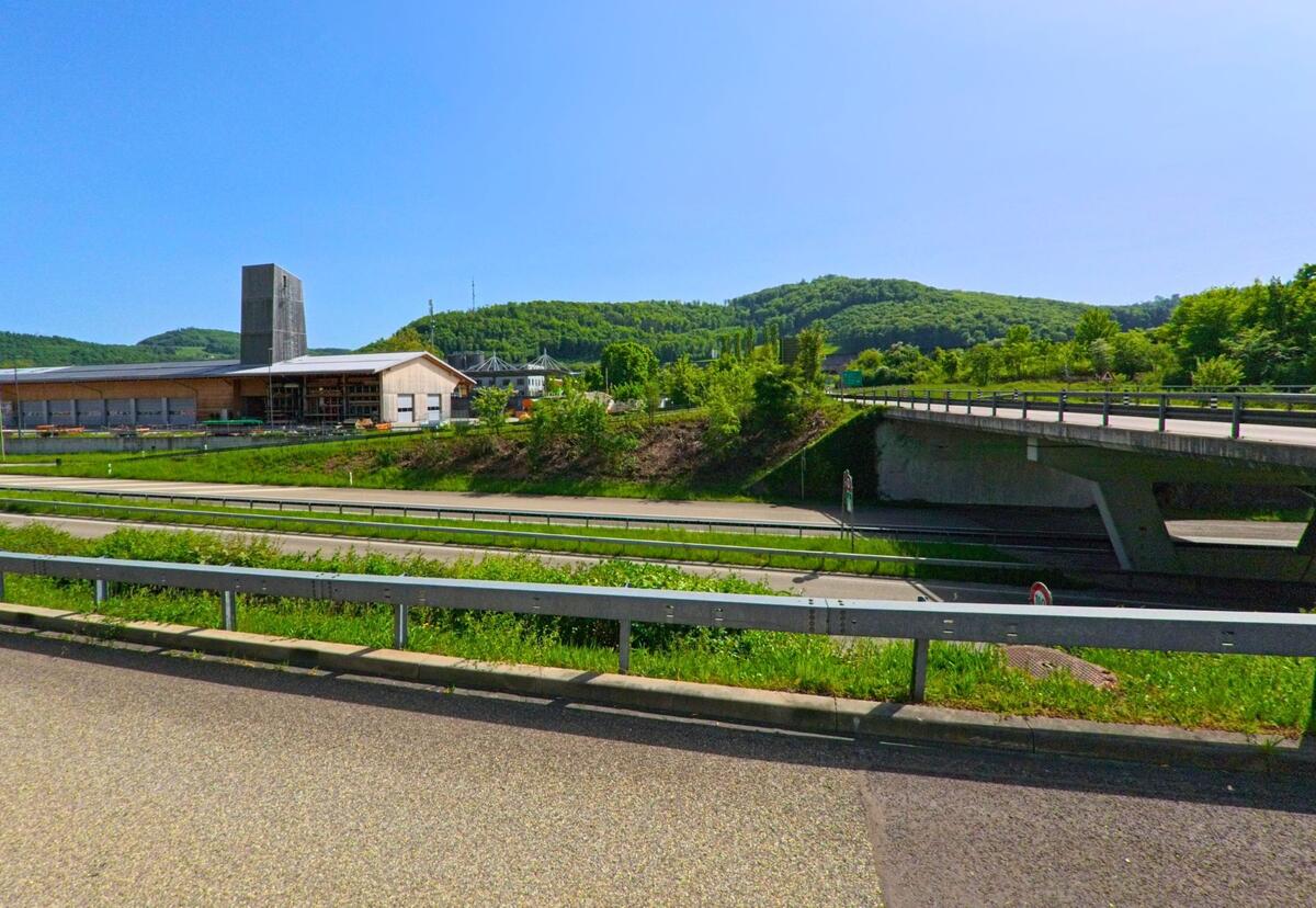 Am Rand von Sissach (4450, Kanton Baselland) verlaeuft die Autobahn neben einem modernen Gewerbebau mit hohem Turm und Blick auf bewaldete Huegel.