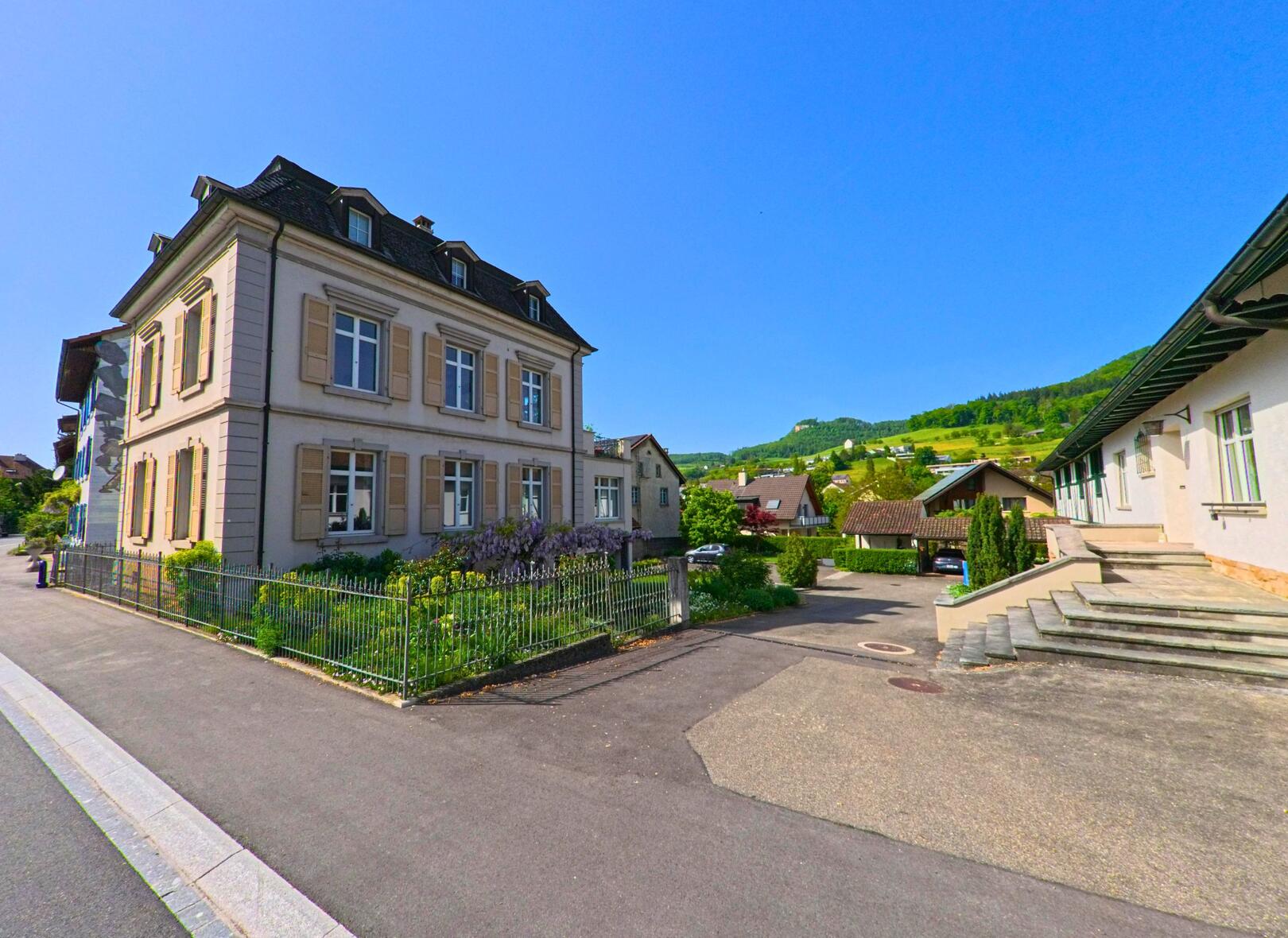 In Sissach (4450, Kanton Baselland) steht ein historisches Gebäude mit eleganten Fensterläden und einem gepflegten Garten, umgeben von modernen Häusern und grünen Hügeln im Hintergrund.
