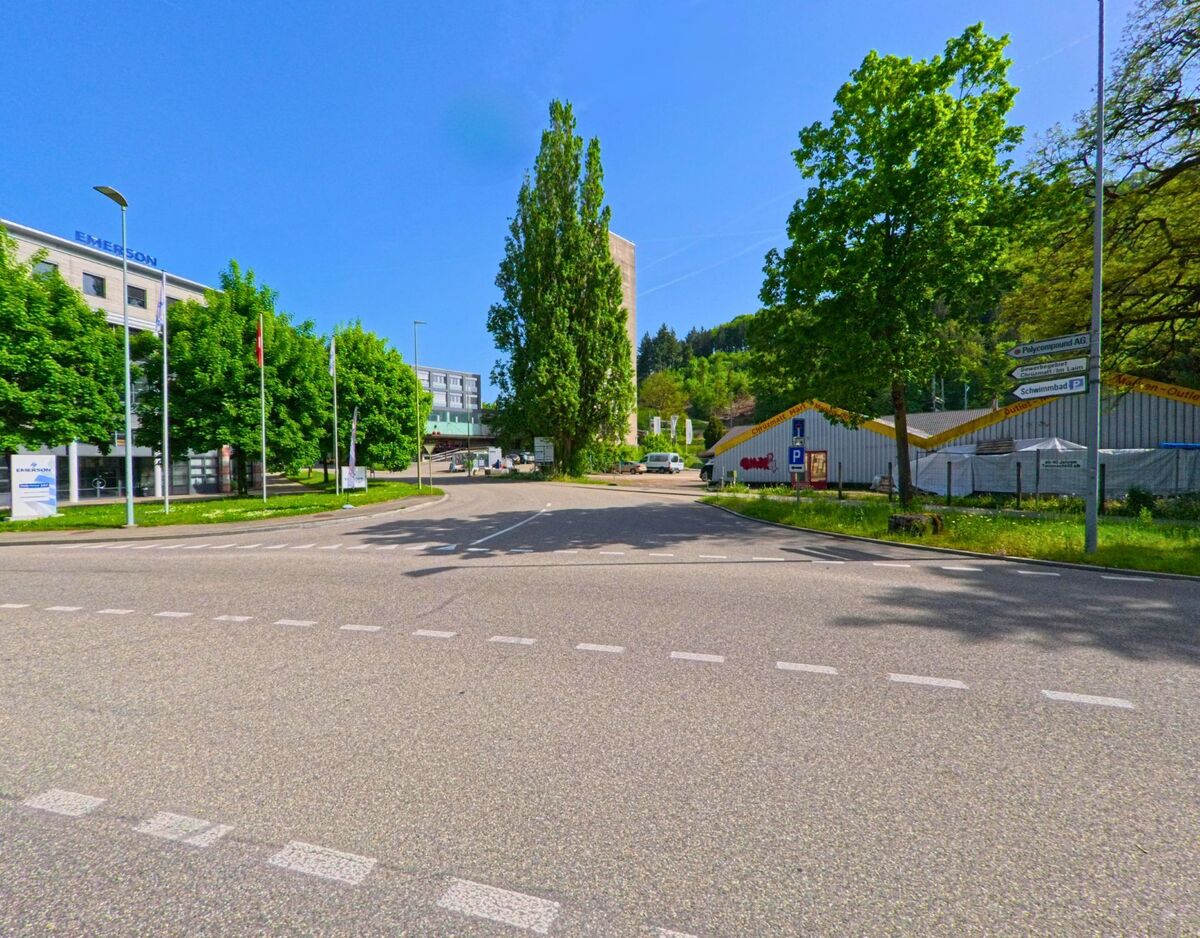 In Sissach (4450, Kanton Baselland) trifft eine breite Kreuzung auf ein Gewerbegebiet mit Buerohaeusern und Lagerhallen am Waldrand.