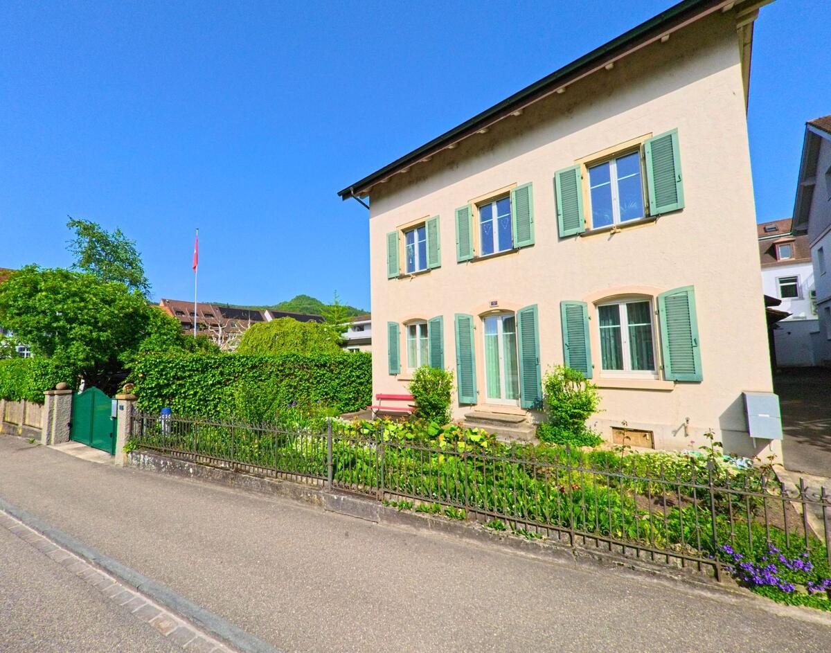 In Sissach (4450, Kanton Baselland) steht ein helles Wohnhaus mit grünen Fensterlaeden und Vorgarten an einer schmalen Quartierstrasse.