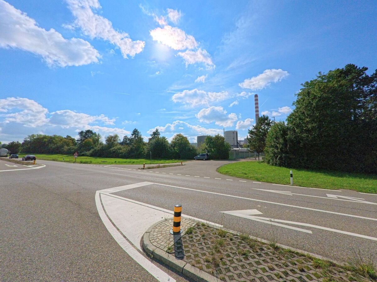 Am Ortsrand von Sisseln (4334, Kanton Aargau) fuehrt eine breite Ausfallstrasse an Wiesen und Baumgruppen vorbei, waehrend im Hintergrund die Industrieanlage mit hohem Schornstein sichtbar ist.