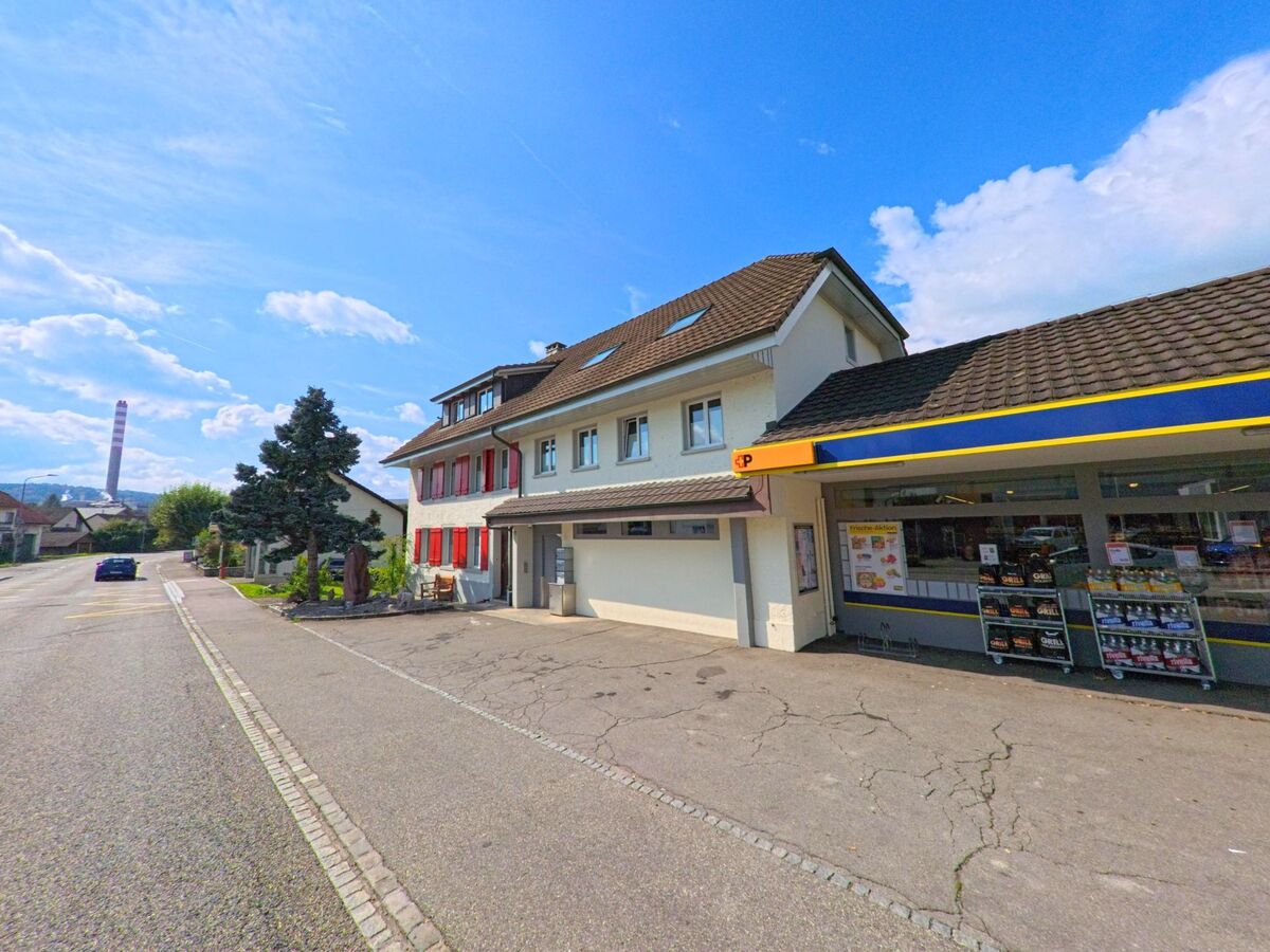 Entlang der Hauptstrasse in Sisseln (4334, Kanton Aargau) stehen ein kleines Geschaeft und ein Wohnhaus mit roten Fensterlaeden, waehrend im Hintergrund der hohe Industrieschornstein sichtbar ist.