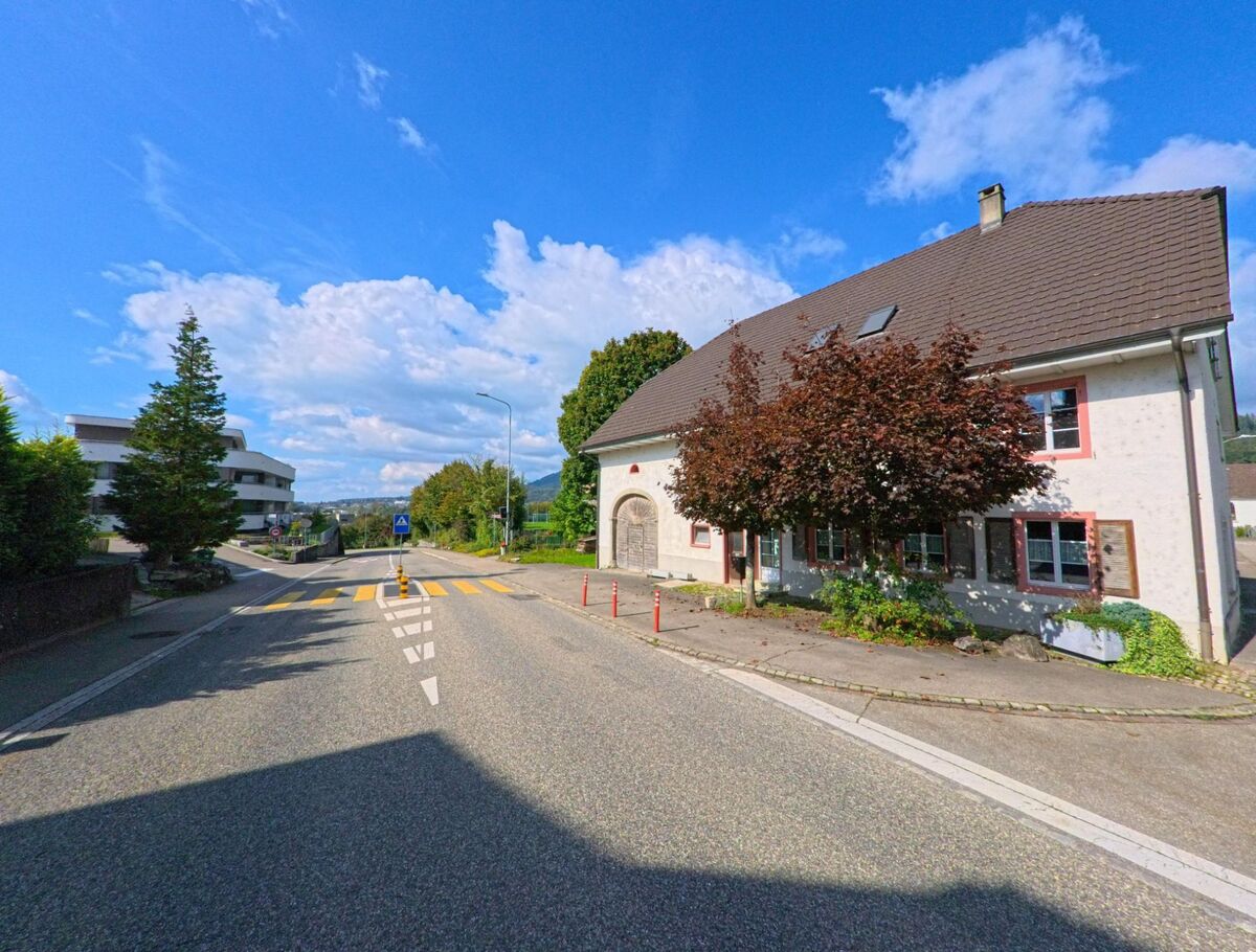 In Sisseln (4334, Kanton Aargau) fuehrt eine leicht abschuessige Strasse mit Fussgaengerstreifen an einem historischen Wohngebaeude mit rotem Laubbaum vorbei, waehrend moderne Bauten im Hintergrund sichtbar sind.