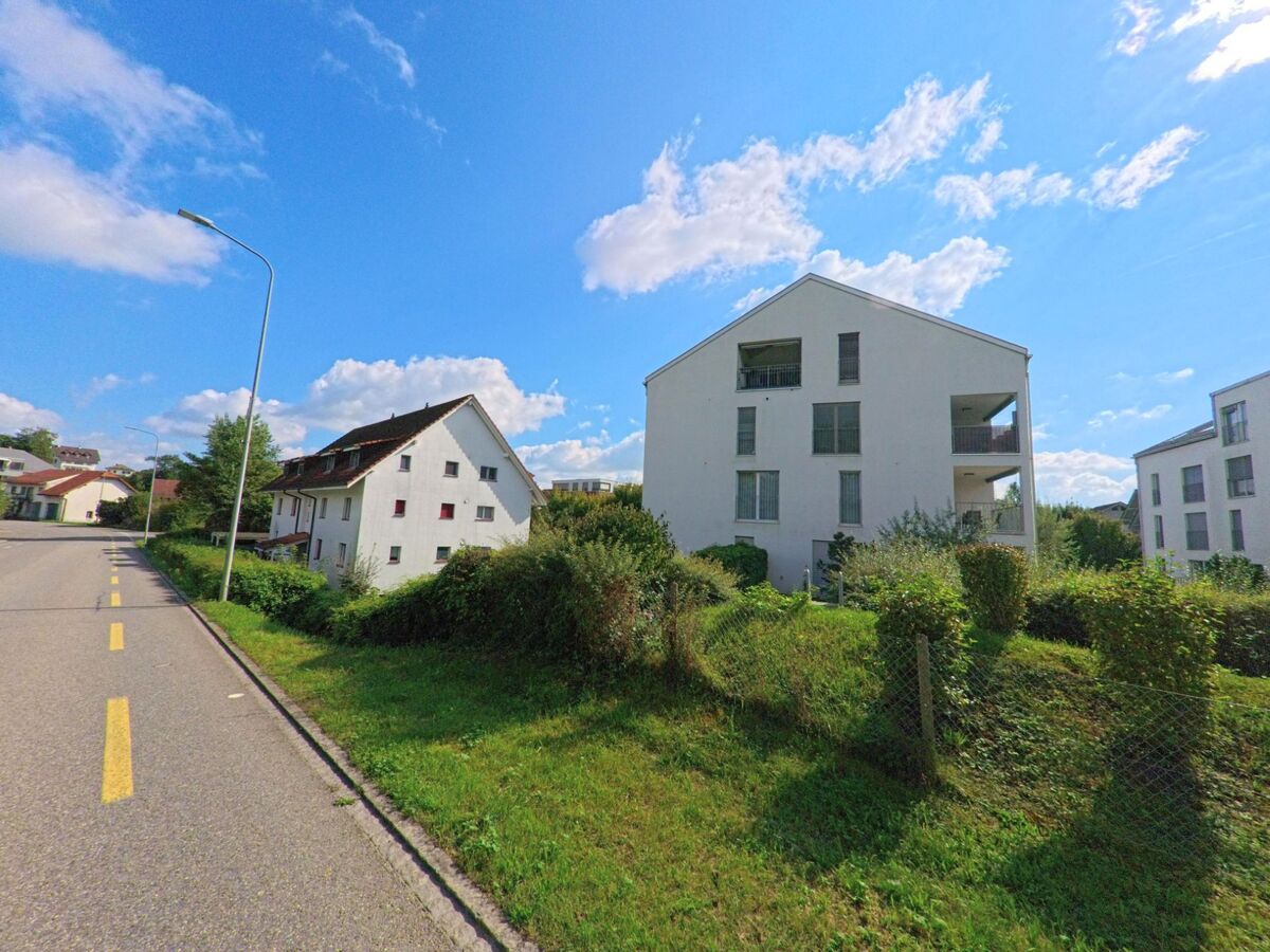 Entlang einer leicht ansteigenden Strasse in Sisseln (4334, Kanton Aargau) stehen moderne Mehrfamilienhaeuser und ein aelteres Wohnhaus, umgeben von Wiesen und Hecken.