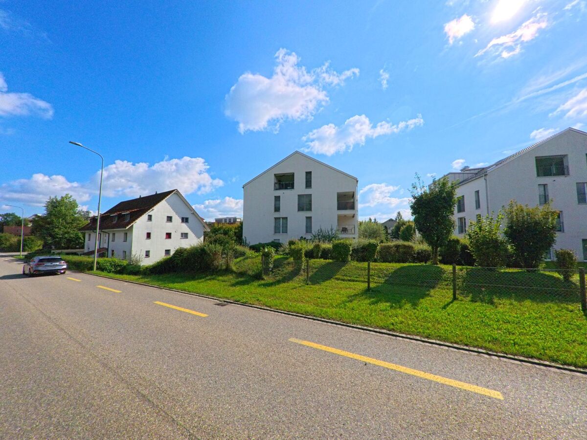 An einer sonnigen Strasse in Sisseln (4334, Kanton Aargau) stehen moderne Mehrfamilienhaeuser neben einem aelteren weissen Wohnhaus, umgeben von gepflegten Gruenflaechen und Hecken.