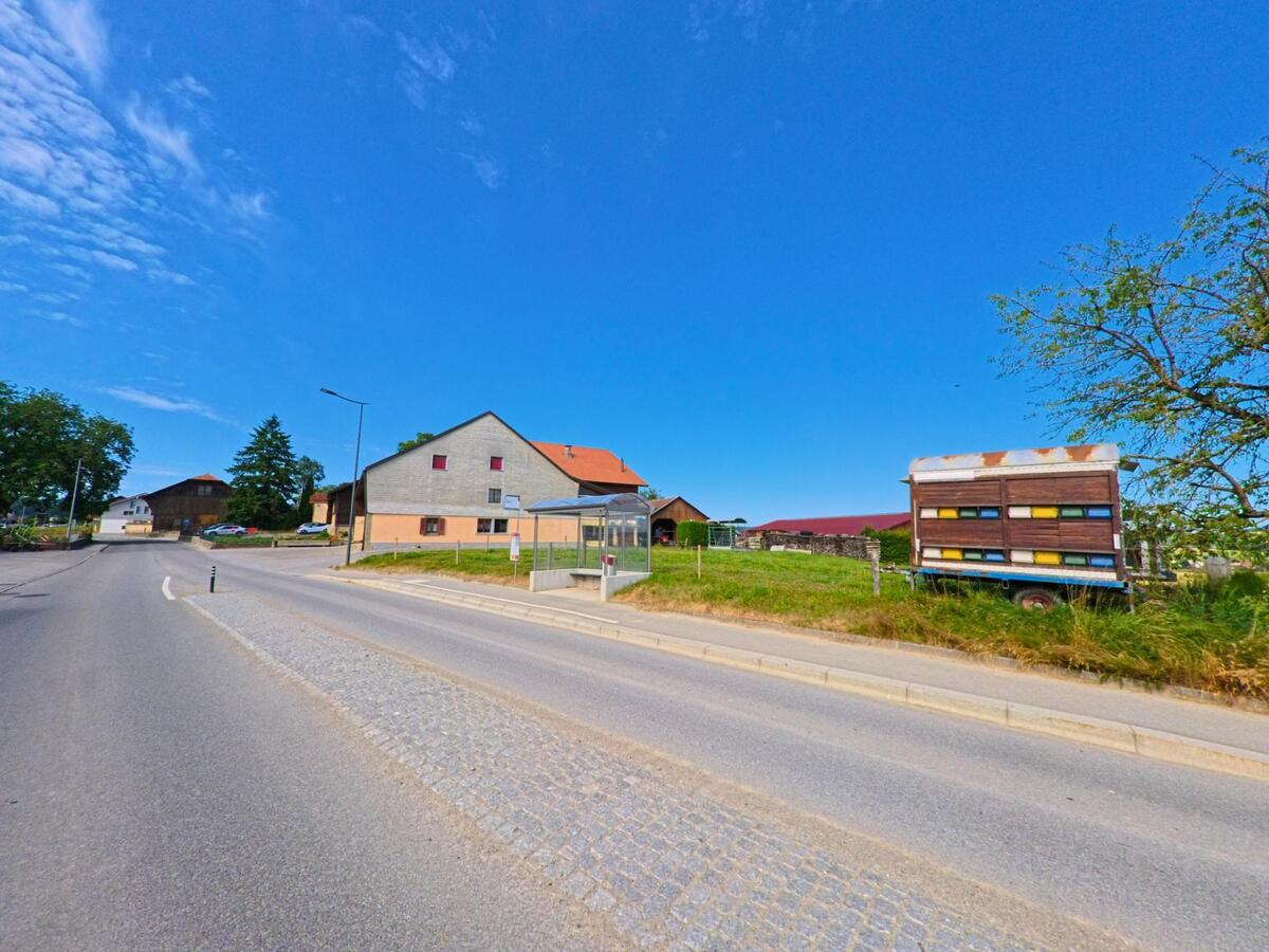 Am Strassenrand in Siviriez (1676, Kanton Freiburg) steht ein mobiler Bienenwagen vor einer Wiese, während gegenüberliegend ein Bauernhof mit Bushaltestelle zu sehen ist.