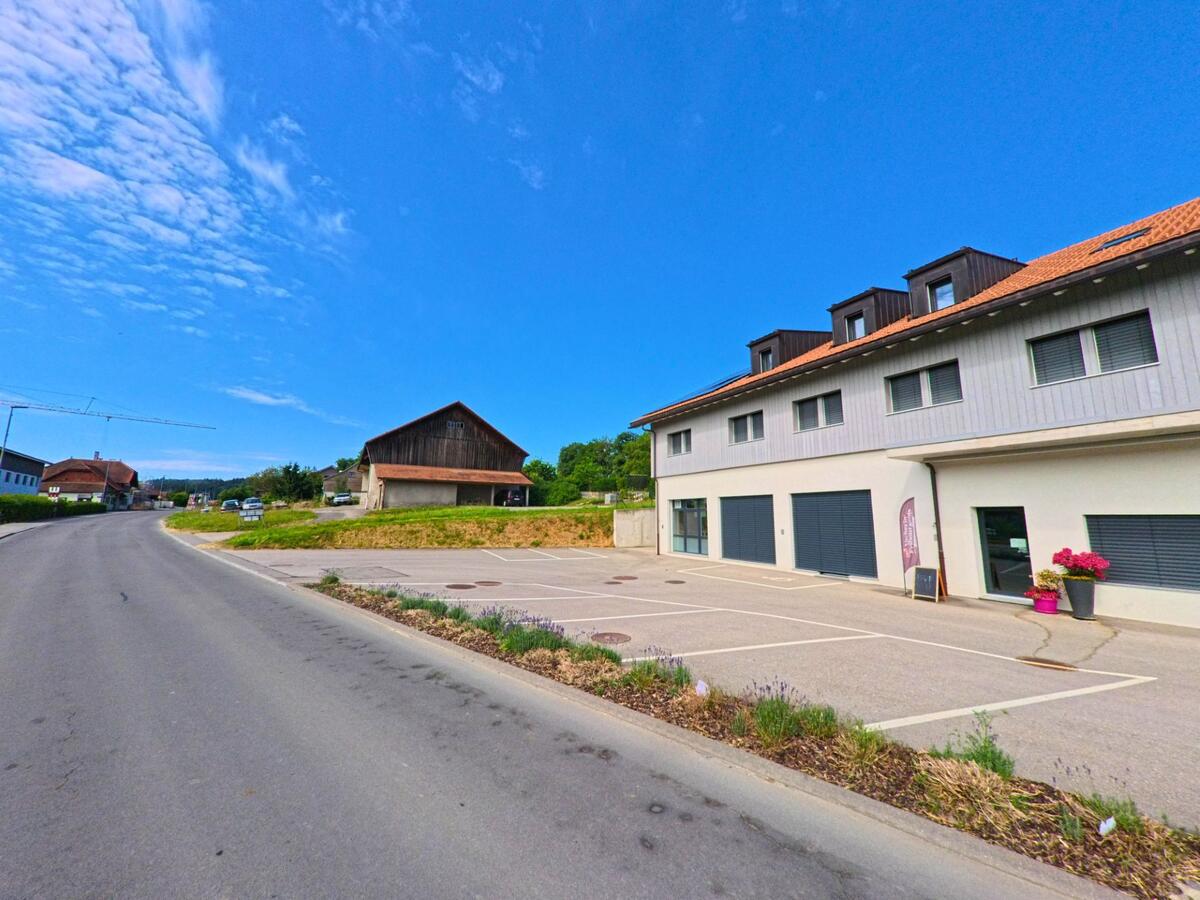 In Siviriez (PLZ 1676, Kanton Freiburg) liegt ein modernes Gebäude mit grossem Parkplatz direkt an der Strasse, gegenüber einem traditionellen Holzstadel.