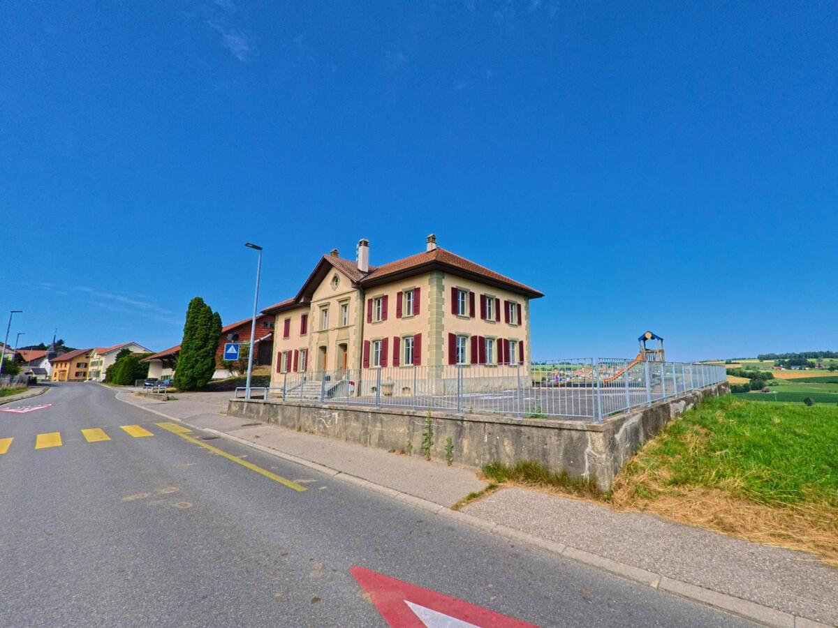 In Siviriez (PLZ 1676, Kanton Freiburg) steht ein klassisches Gebäude mit roten Fensterläden und vorgelagertem Spielplatz oberhalb einer leicht erhöhten Mauer am Strassenrand.