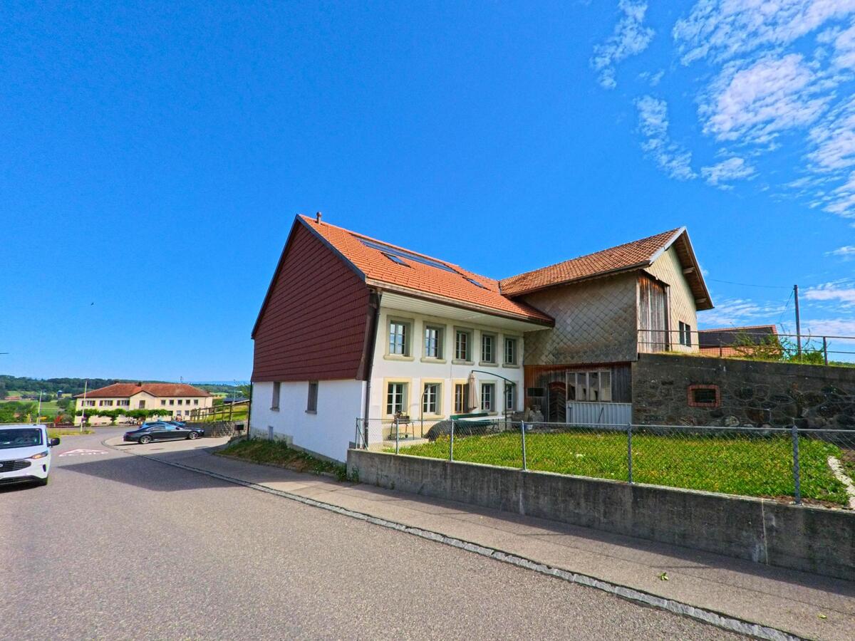 In Siviriez (PLZ 1679, Kanton Freiburg) steht ein traditionelles Wohnhaus mit angebautem Scheunenteil und eingezäuntem Vorgarten an einer leicht ansteigenden Strasse.