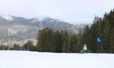 In Neuenkirch (6206, Kanton Luzern) faehrt ein Skilaeufer durch einen Torlauf auf der Piste, waehrend sich im Hintergrund ein Regenbogen ueber die bewaldeten Haenge spannt.