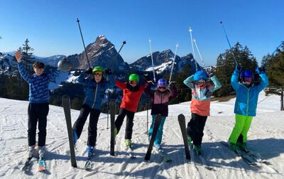 In Lauerz (6424, Kanton Schwyz) stehen junge Skifahrerinnen und Skifahrer lachend auf einer sonnigen Piste, mit erhobenen Stoecken und markanter Bergkulisse im Hintergrund.