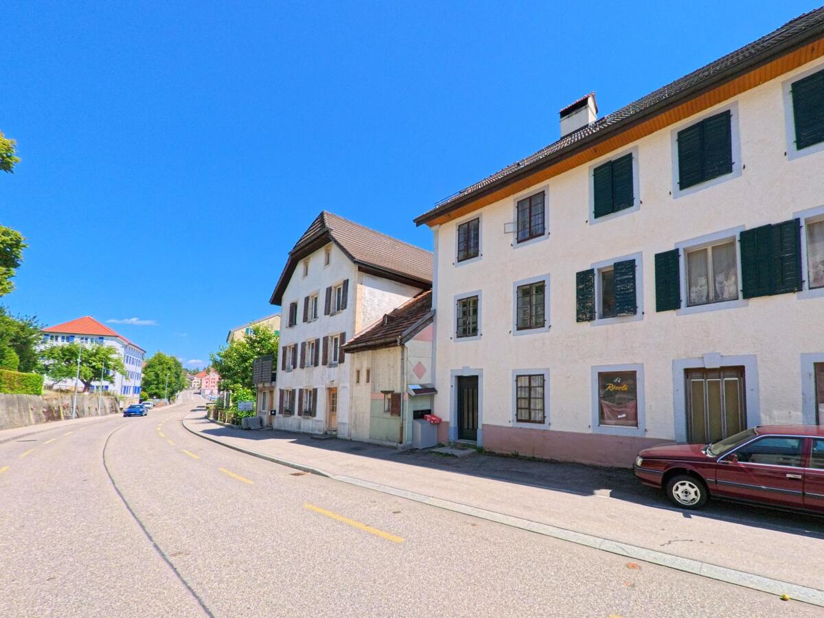 Entlang einer geschwungenen Strasse in Sonvilier (2615, Kanton Bern) stehen mehrstöckige Wohnhaeuser mit unterschiedlich gefaerbten Fensterlaeden und ein geparktes Auto.