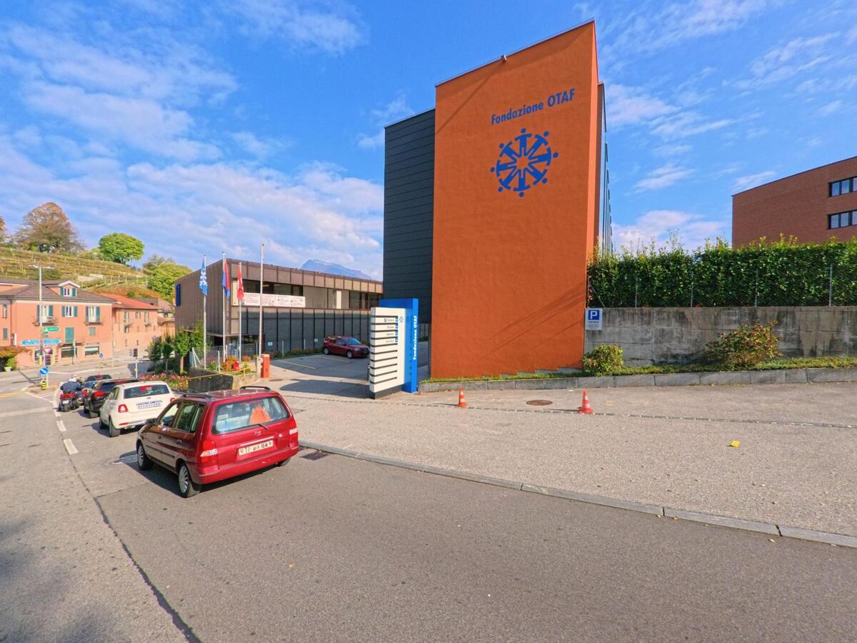 An einer Nebenstrasse in Sorengo (6924, Kanton Tessin) steht ein markantes Gebäude der Fondazione OTAF neben parkierenden Autos und weiteren Bauten.