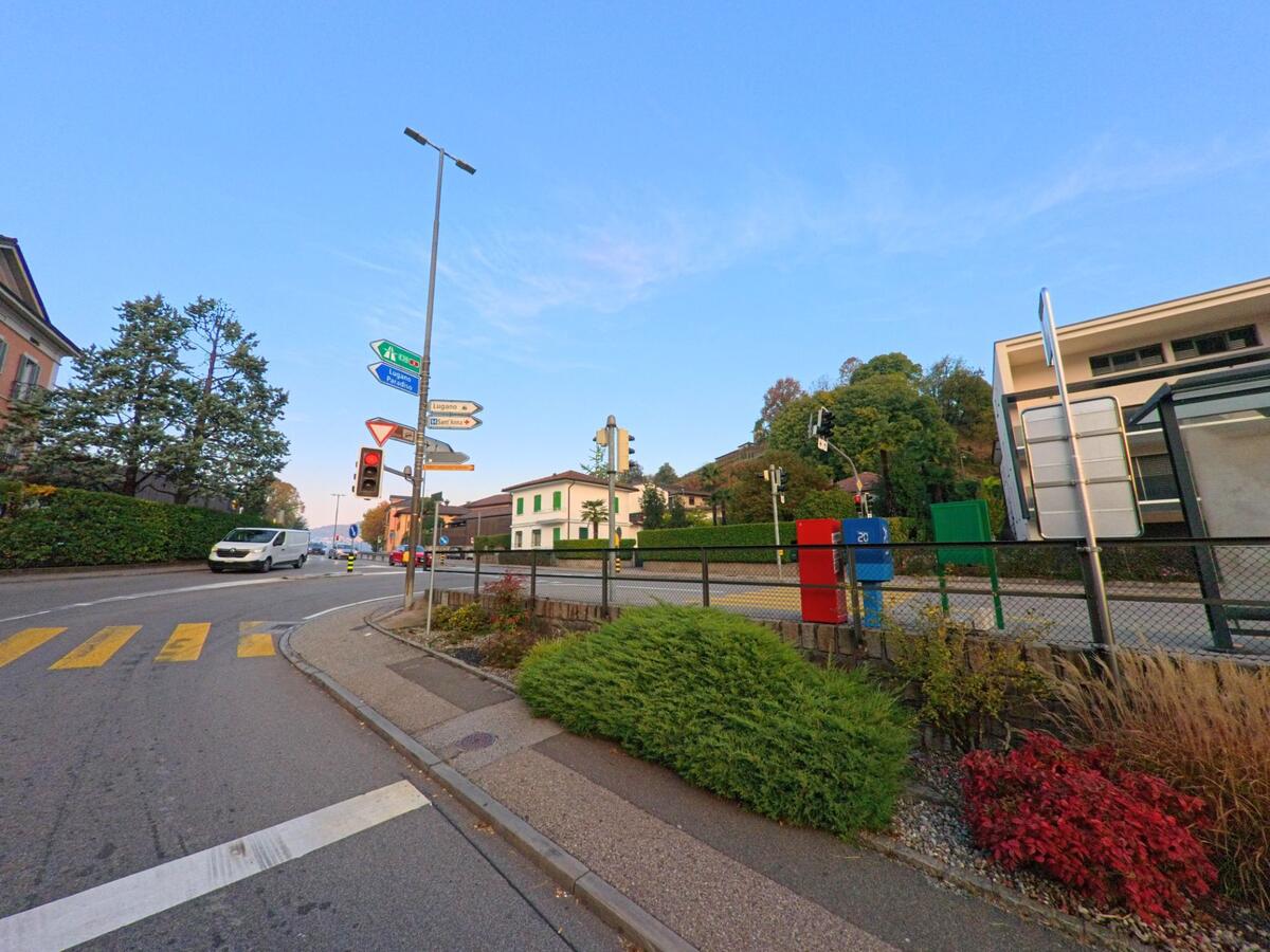 An einer signalgeregelten Kreuzung in Sorengo (6924, Kanton Tessin) stehen Wegweiser, Ampeln und Briefkästen entlang der Strasse vor begrünten Hängen.