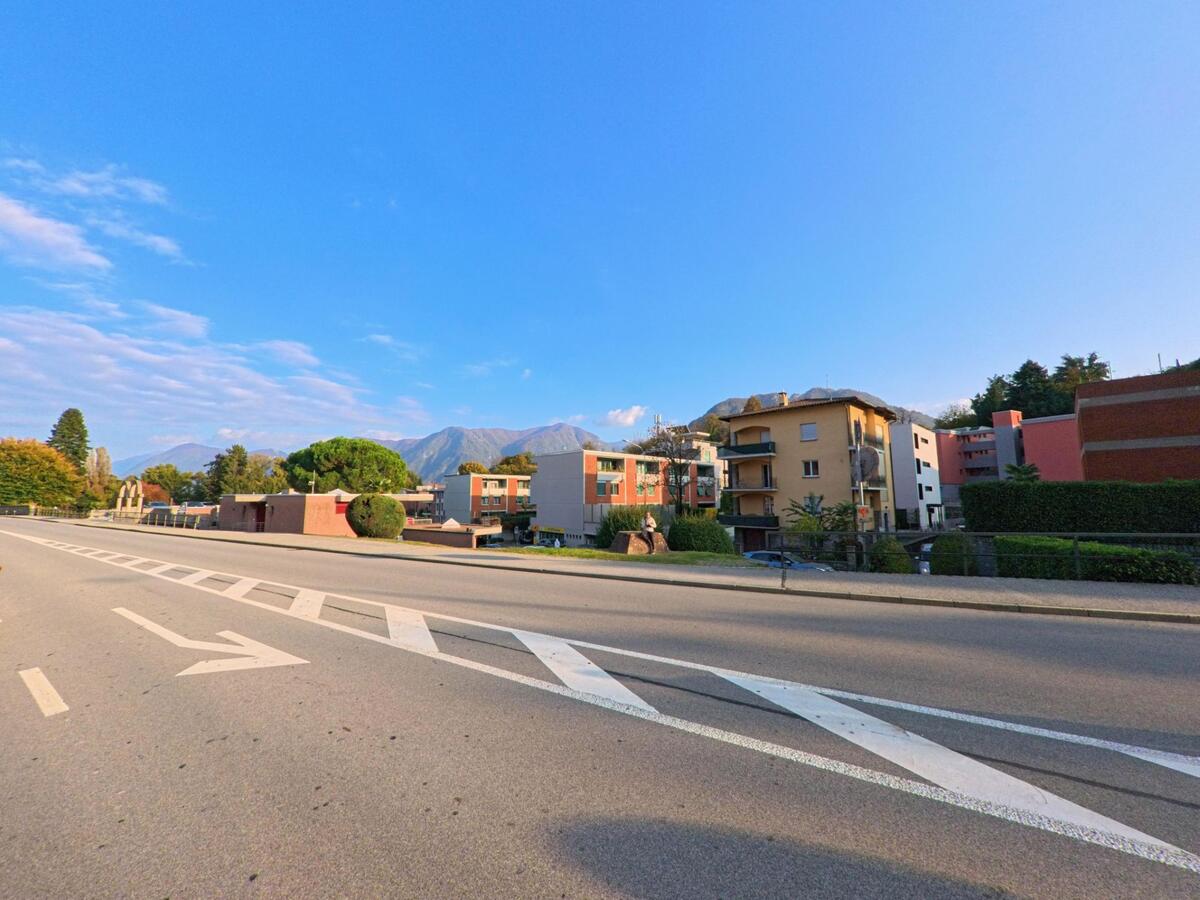 Über eine breite Fahrbahn hinweg öffnet sich in Sorengo (6924, Kanton Tessin) der Blick auf Wohnbauten mit dahinterliegenden Hügeln und Bergen unter klarem Himmel.