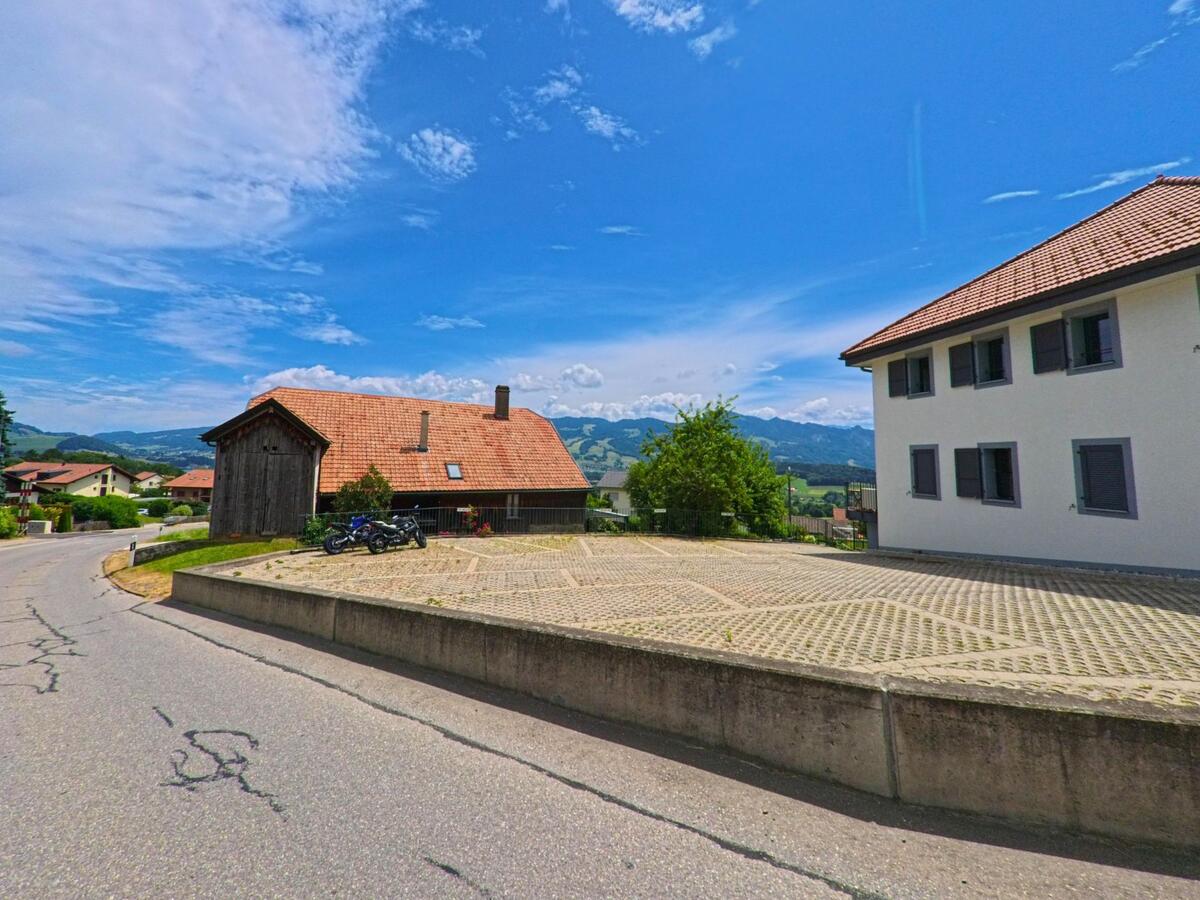 In Sorens (1642, Kanton Freiburg) treffen ein renoviertes Bauernhaus mit Holzfassade und ein modernes Wohnhaus an einer kurvigen Strasse aufeinander, im Hintergrund erstrecken sich die Alpen.
