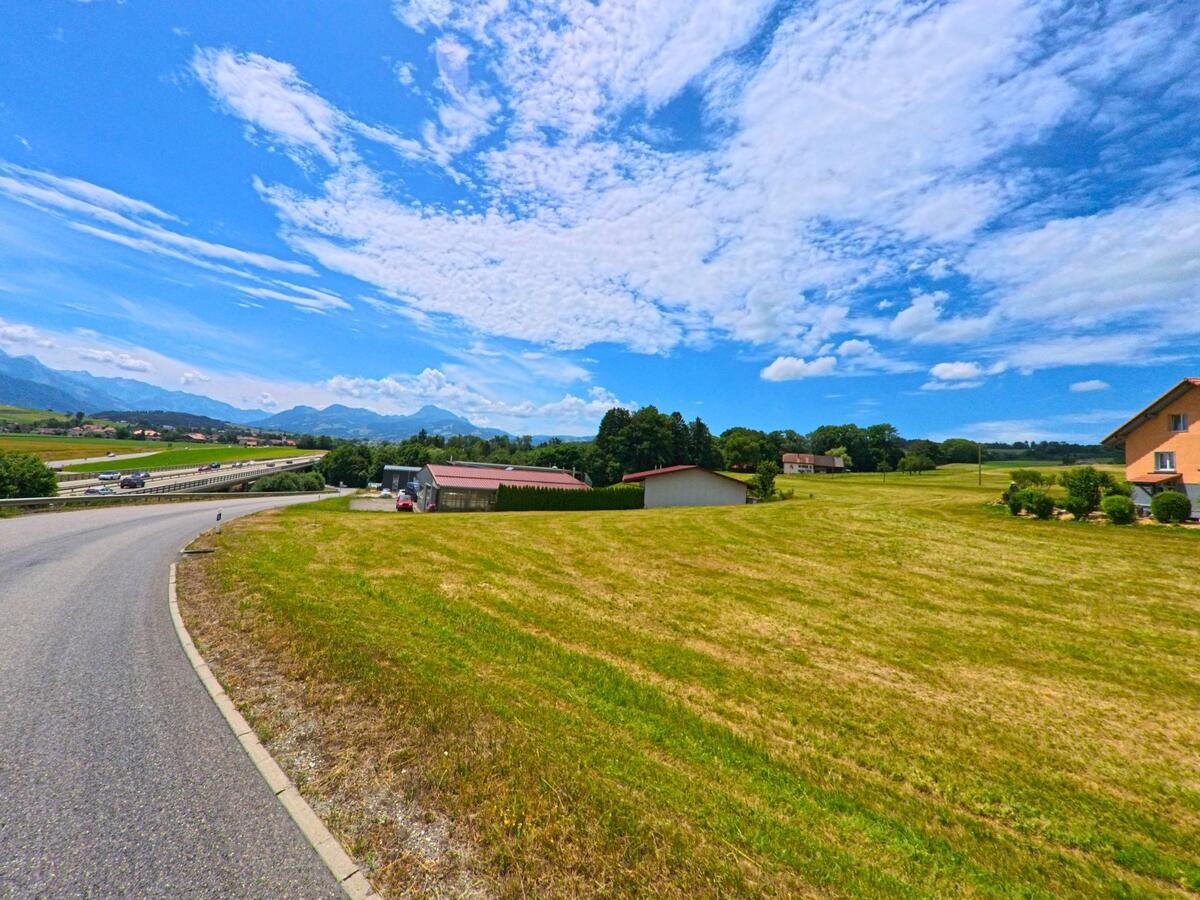 Eine geschwungene Strasse führt in Sorens (1642, Kanton Freiburg) an Wiesen vorbei in Richtung Autobahn, während im Hintergrund die Berge unter einem weit geöffneten Himmel sichtbar sind.
