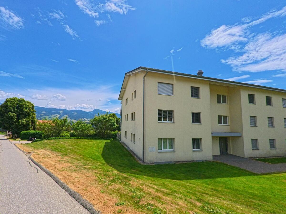 Ein helles Mehrfamilienhaus in Sorens (1642, Kanton Freiburg) steht an einer ruhigen Strasse mit gepflegtem Rasen und Blick auf die bewaldeten Hügel des Umlands.
