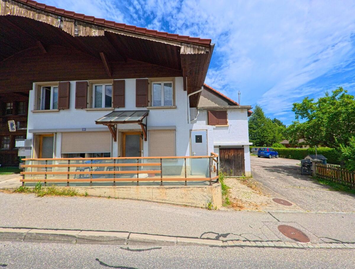 In Sorens (1642, Kanton Freiburg) steht ein Wohnhaus mit braunen Fensterläden und Glasvordach direkt an der Strasse, daneben verläuft ein kleiner Weg mit parkendem Auto.