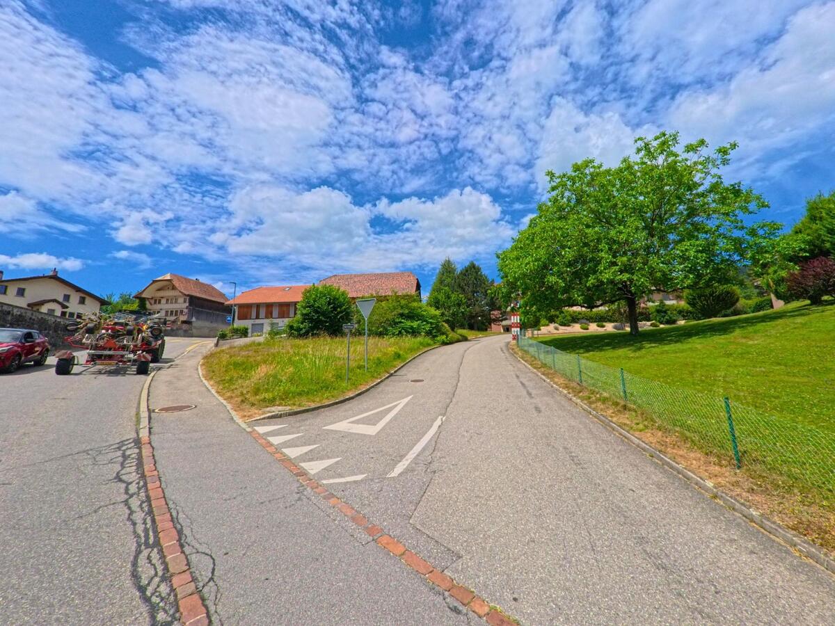 In Sorens (1642, Kanton Freiburg) teilt sich die Strasse an einer Gabelung mit Blick auf ein Bauernhaus, während ein Baum auf der rechten Seite eine gepflegte Grünfläche beschattet.