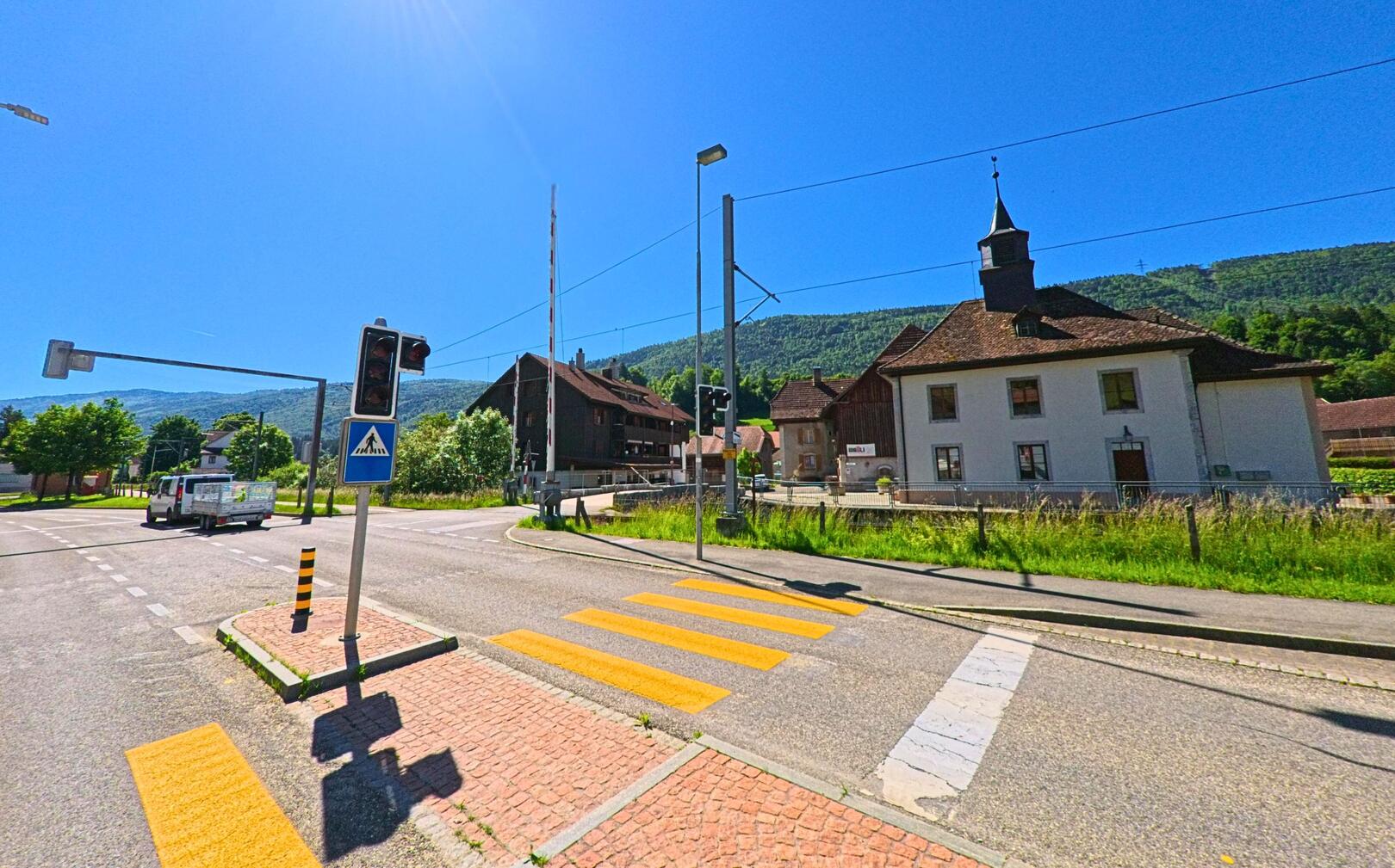 In Sorvilier (PLZ 2736, Kanton Bern) kreuzt eine Verkehrsinsel mit Fussgängerstreifen und Ampelanlage den Bahnübergang nahe der Kirche im Dorfzentrum.