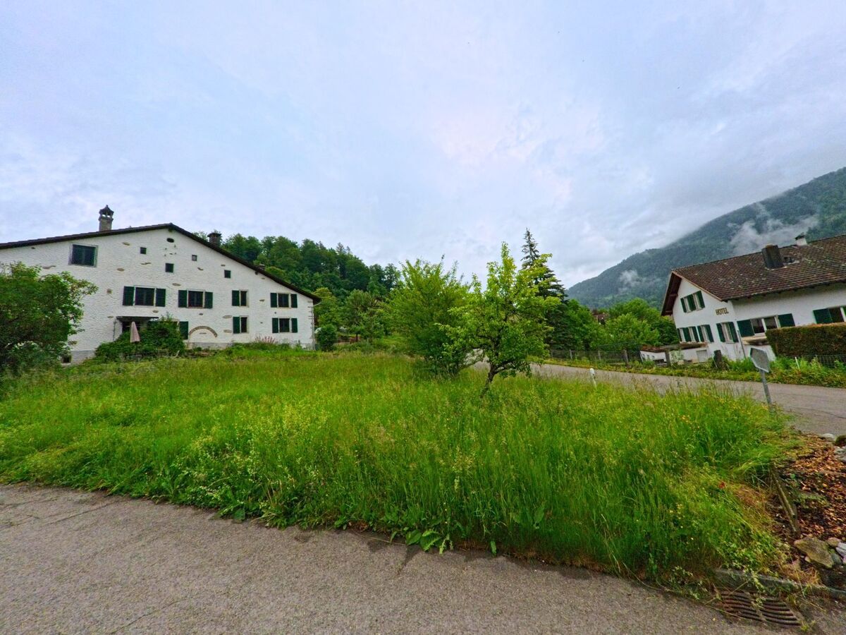 Zwei weiss verputzte Gebaeude mit gruenen Fensterlaeden stehen in Soubey (2887, Kanton Jura) gegenueber einer Grasflaeche am Strassenrand, umgeben von bewaldeten Huegeln und Nebelschwaden.