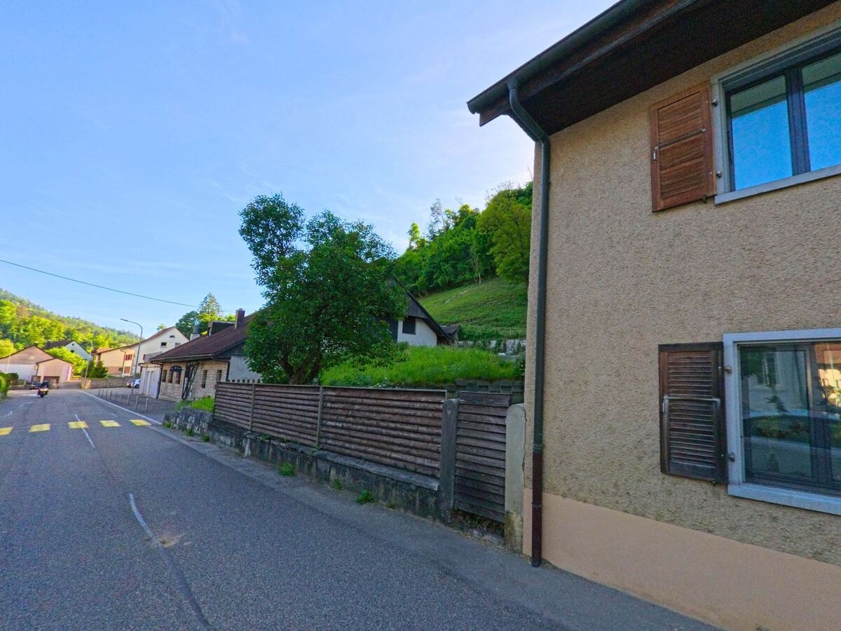 In Soyhières (2805, Kanton Jura) verläuft eine Hauptstrasse mit Fussgängerstreifen entlang eines Hangs, flankiert von Wohnhäusern mit Fensterläden und Gartenmauern.
