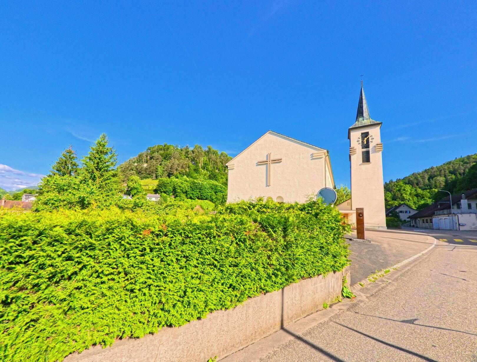 In Soyhières (2805, Kanton Jura) steht eine helle Kirche mit markantem Glockenturm an einer Strassenkurve, umgeben von grün bewachsenen Hügeln.